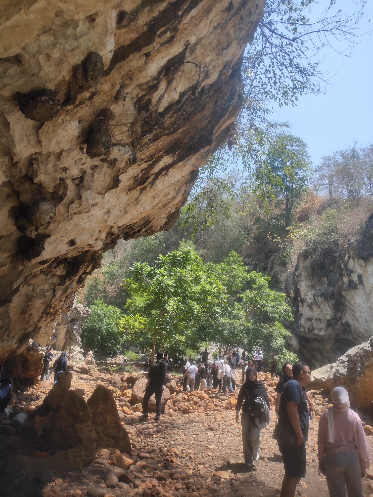 A group of people are walking on a rocky path leading into a cave at the Gua Arca archaeological site in Cipatat, Indonesia. The cave has a large, rough rock overhang, and the surrounding environment is lush with green trees and rocky hills under a clear blue sky. The people are dressed casually and appear to be tourists or visitors exploring the site. Some are walking ahead, while others are pausing to observe the surroundings or take photos. The ground is covered in small and medium-sized rocks, indicating a natural, undeveloped terrain.