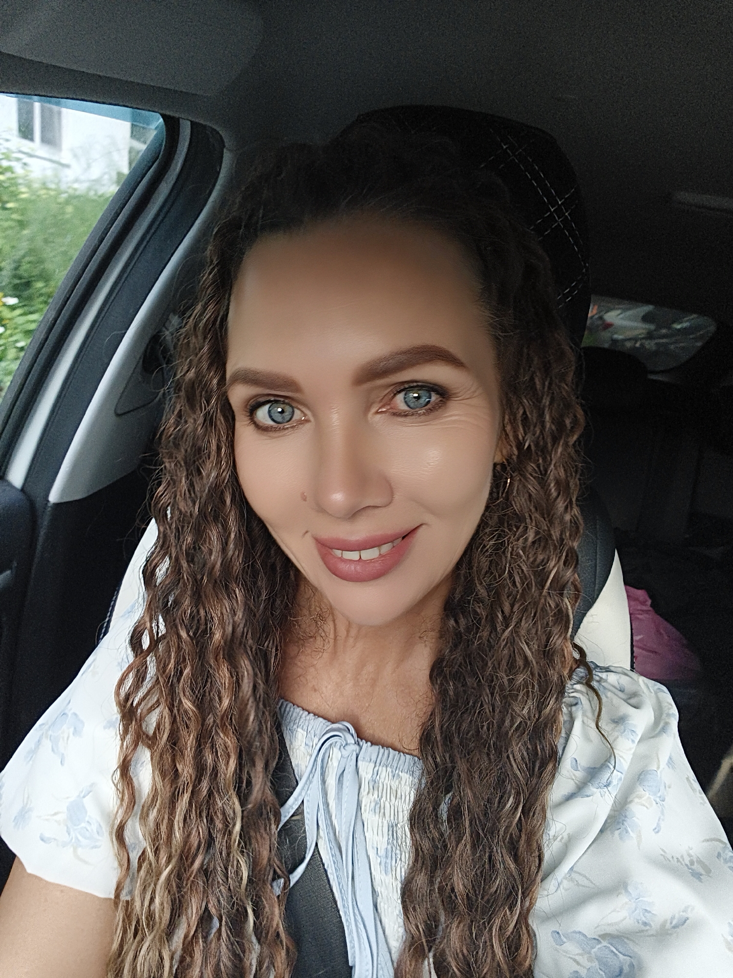 A woman with long, curly brown hair and blue eyes is taking a selfie in a car. She is wearing a light blue and white floral print top. The background shows the interior of the car and greenery outside the window. The woman appears to be in a relaxed setting, possibly on a trip or commute. The image was taken in Chaikovsky, Russia.