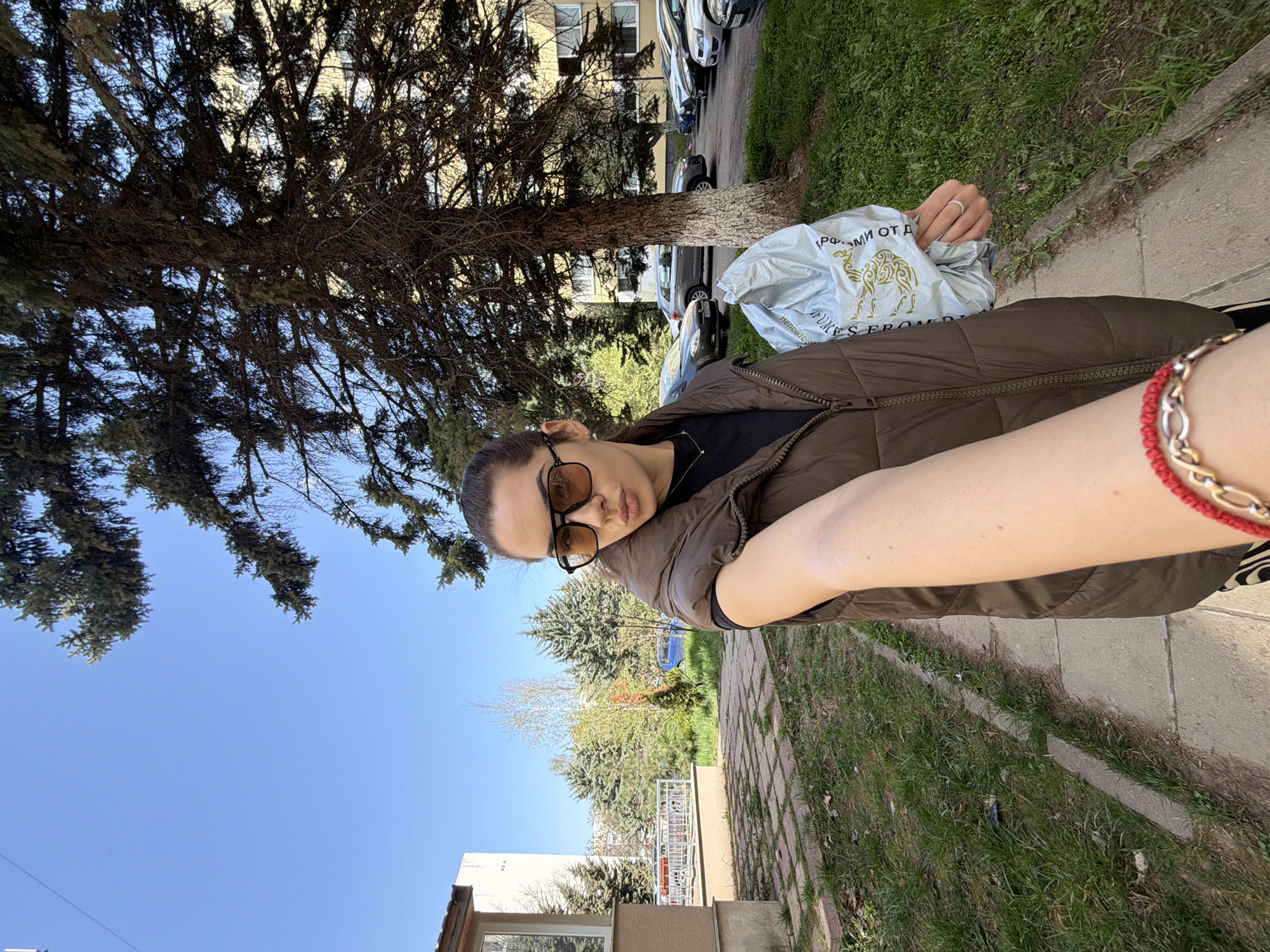 The image is a selfie-style shot of a young woman outdoors in Sofia, Bulgaria, on a clear, sunny day. The woman, seen from the chest up, has her dark hair pulled back and wears black-framed sunglasses with amber lenses, which obscure her eyes. Her expression is neutral to slightly pouty. She is dressed in a dark brown puffer vest over a black top, and a subtle silver-colored necklace is visible. On her left ring finger, she wears a simple silver-colored ring, and on her right wrist, a red braided bracelet with a gold-colored chain link. She holds a light-colored plastic bag in her left hand, which features a prominent camel graphic. Partially visible Cyrillic text on the bag includes "Р. ФОМИИ ОТ Д..." and "Неповто...", along with an English text fragment "...KES FROM D...".
The background reveals an urban setting with a paved sidewalk and a grassy strip bordered by mature trees, including conifers. The bright sun casts distinct shadows, indicating strong daylight. Behind the trees and a line of parked cars, an apartment building with multiple windows is visible. The sky is a vibrant, cloudless blue, signifying excellent weather. The scene suggests a casual walk or outing in a residential area of Sofia.