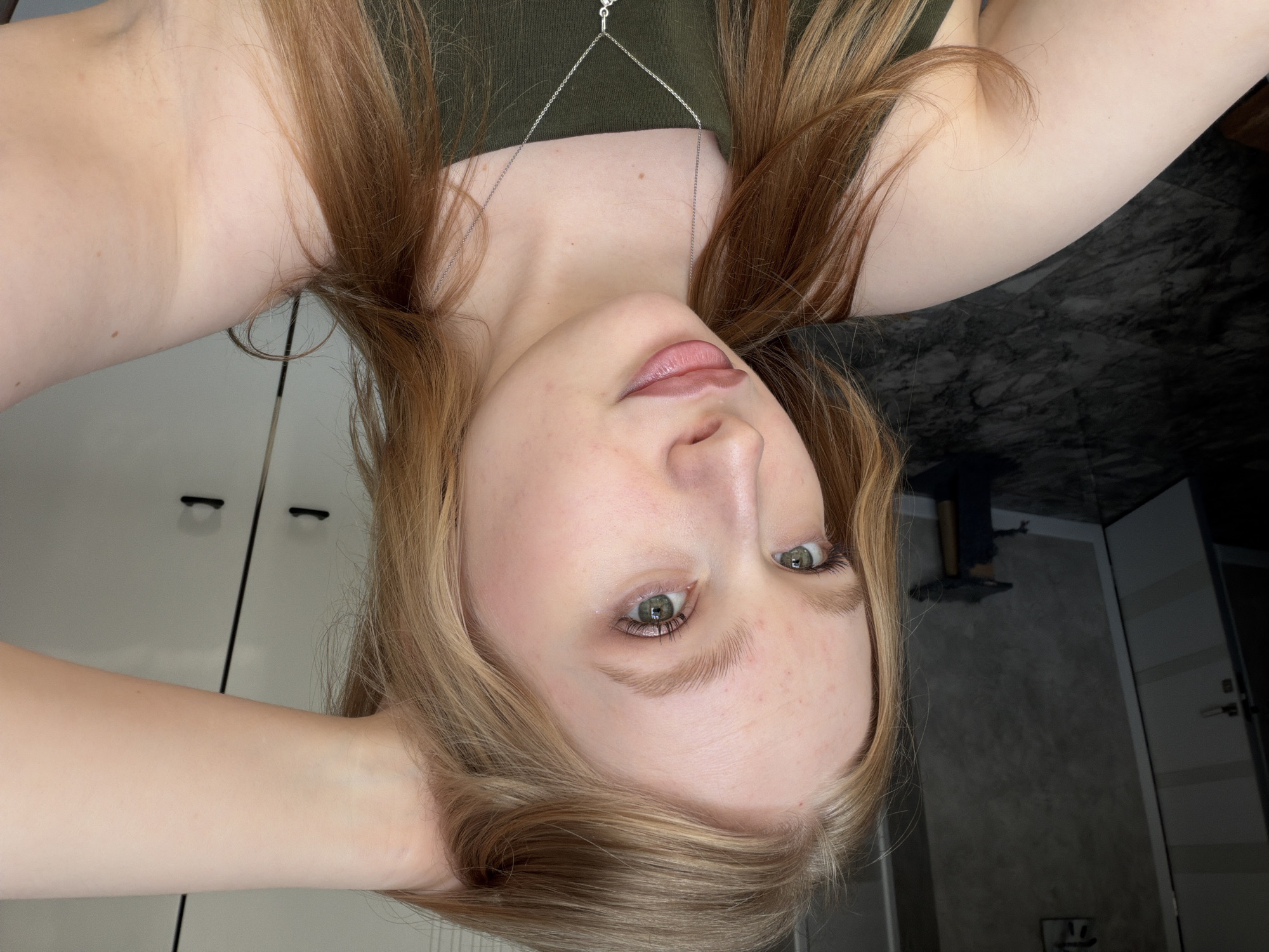 A pale-skinned person with light brown and blonde hair is posing for a selfie, their head tilted back. They are wearing a green top and a silver necklace. Their eyes are green, and their lips are a light pink. The background appears to be a room with white cabinets and a dark marble-like surface. The person is likely located in Rossiyskiy, Russia, based on the request. No other details are discernible about the location.