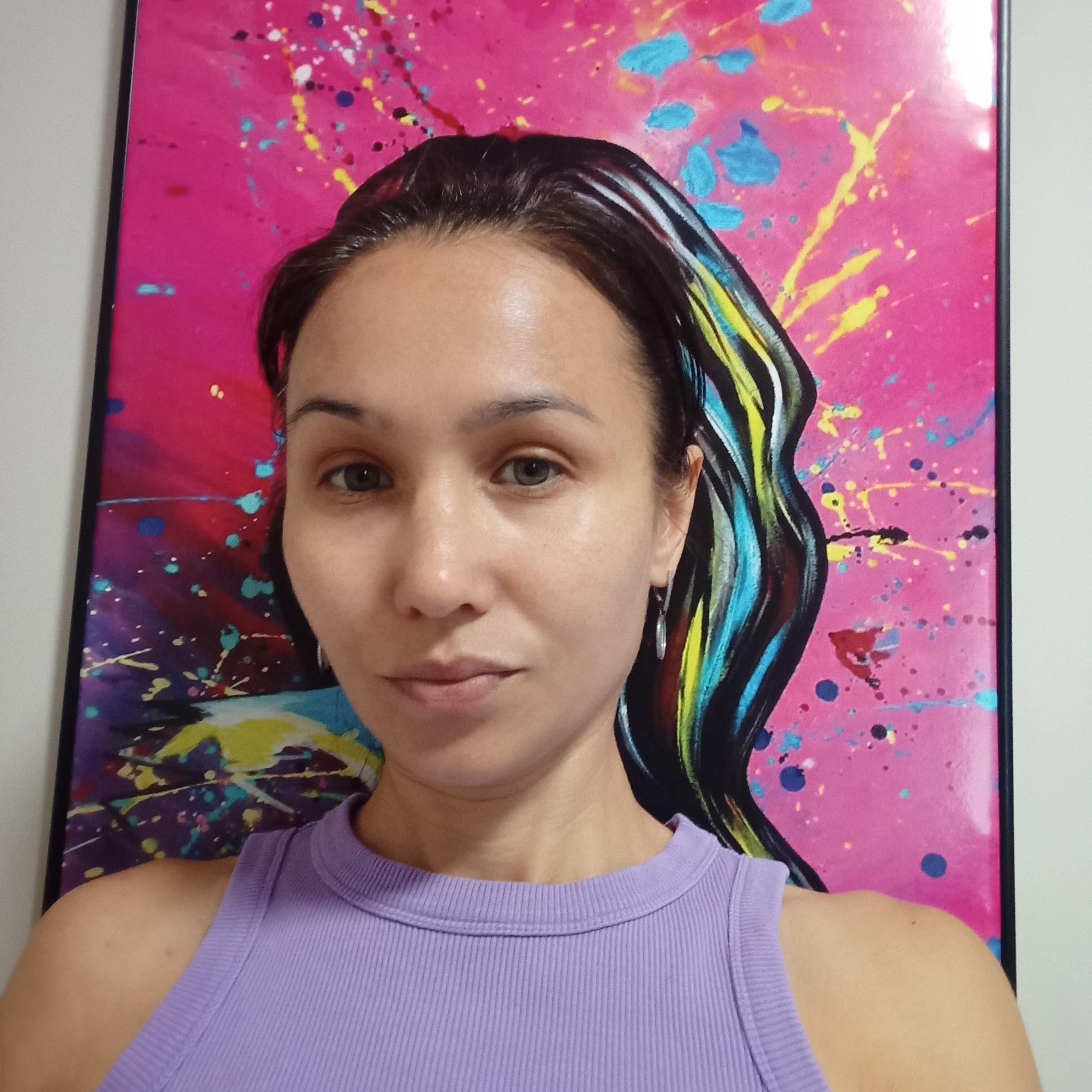 A woman is posing for a selfie in front of a vibrant, abstract painting. The painting features a dominant pink background with splatters of various colors, including yellow, blue, and white, creating a dynamic and energetic effect. A stylized black silhouette with streaks of bright blue, yellow, and white runs down the right side of the painting, suggesting a figure or hair.
The woman has dark hair pulled back, revealing her face. She is wearing a ribbed, lavender tank top. Her expression is neutral, and she is looking directly at the camera. A small, silver earring is visible on her right ear. The background wall to the left of the painting is a plain white. The lighting appears to be soft and even, illuminating her face without harsh shadows. The overall impression is a casual portrait set against a striking piece of modern art.
The provided information states this image is located in the city of Pervomaysky, Russia.