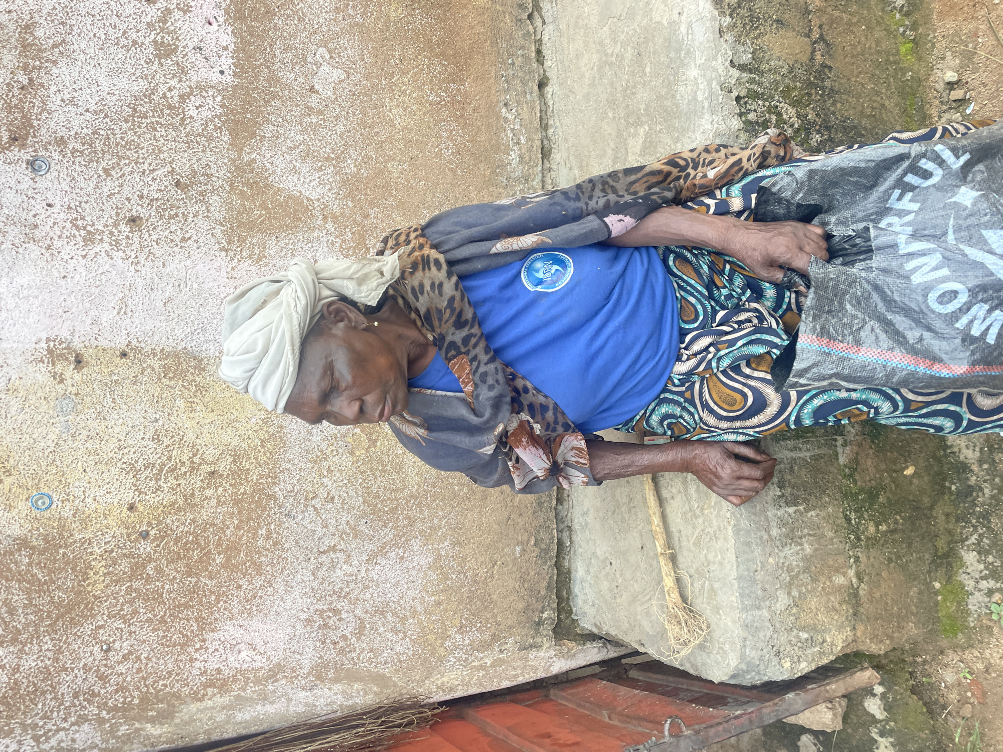 In Jos, Nigeria, an elderly woman with dark skin, wearing a white headscarf, a blue t-shirt with a logo, a patterned skirt, and a leopard-print scarf, sits on concrete steps in front of a textured, weathered wall. She holds a black plastic bag with white text. A broom-like object lies on the step beside her. The wall appears to be made of mud plaster with some staining and debris. The ground is composed of dirt and some patches of moss. The scene suggests an outdoor, possibly informal, setting within the city.