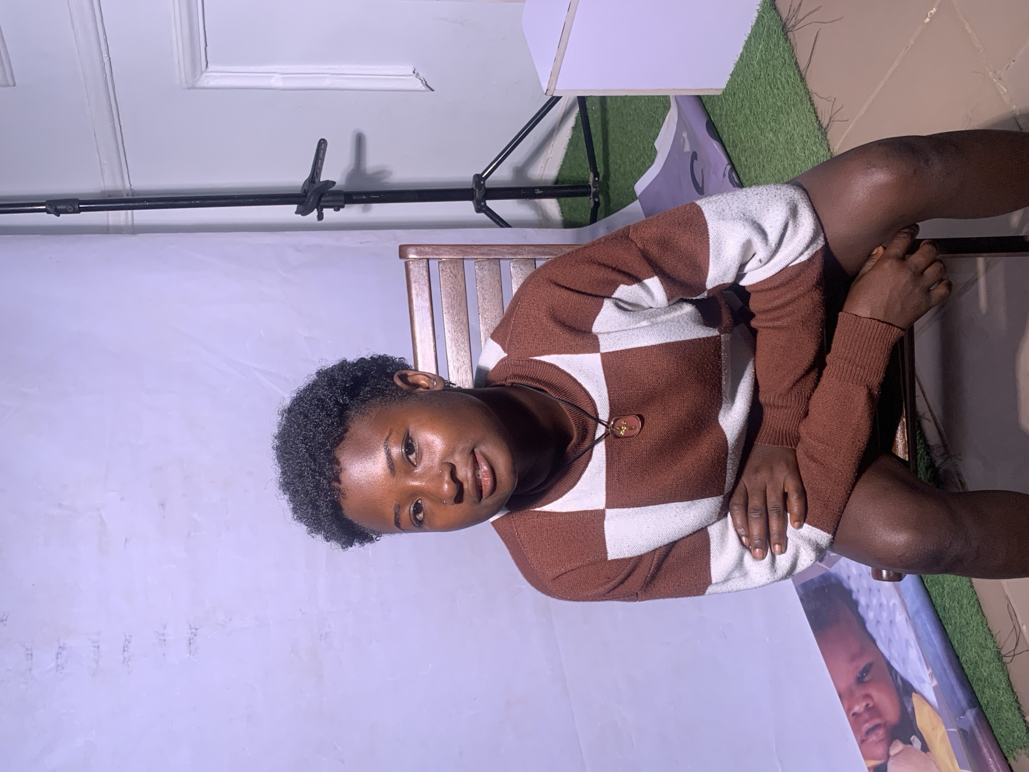 In a studio setting located in Barakin Akawo, Nigeria, a young Black woman is seated on a wooden chair. She has short, dark, curly hair and is wearing a brown and white checkered sweater. Her arms are crossed, and she is looking directly at the camera. Behind her, a white backdrop is visible, along with a lighting stand and a white cube on the right side of the frame. A rolled-up banner featuring a child's face is placed on the floor in front of the backdrop. The floor appears to be covered with green artificial turf in some areas and tiled in others.