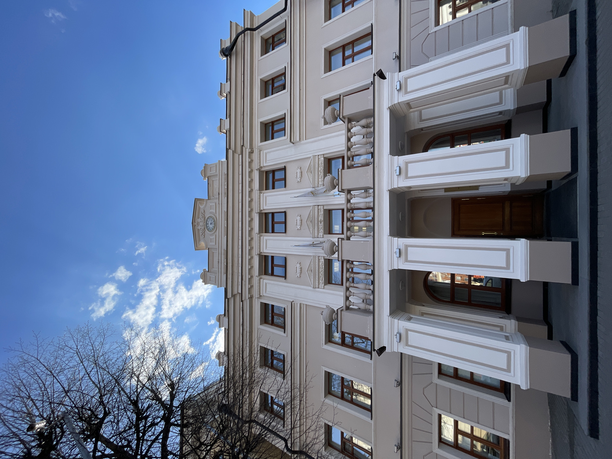 The image captures the exterior of a building in Kazan, Russia. The structure is multi-storied and exhibits architectural elements such as rectangular windows with brown frames, ornate trim details, and projections resembling balconies. The building is beige, with white architectural accents. The sky above is blue with scattered white clouds, and bare branches of trees are visible in the lower left corner of the image. The photograph is taken from a low angle, looking upwards at the building, which appears to be situated on a street.