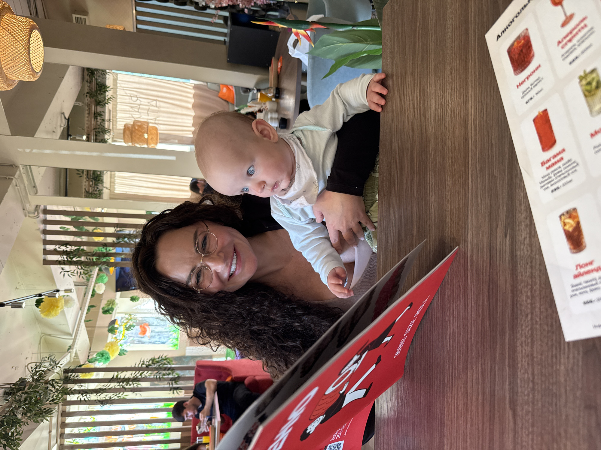 The image depicts a woman holding a baby in a restaurant. The woman is wearing glasses and smiling, while the baby is looking away from the camera. They are seated at a wooden table, with a menu in the foreground. The restaurant appears to be decorated with plants and lights, creating a relaxed atmosphere. In the background, other patrons can be seen. The overall setting suggests a casual dining experience. The location is identified as Sochi, Russia.
