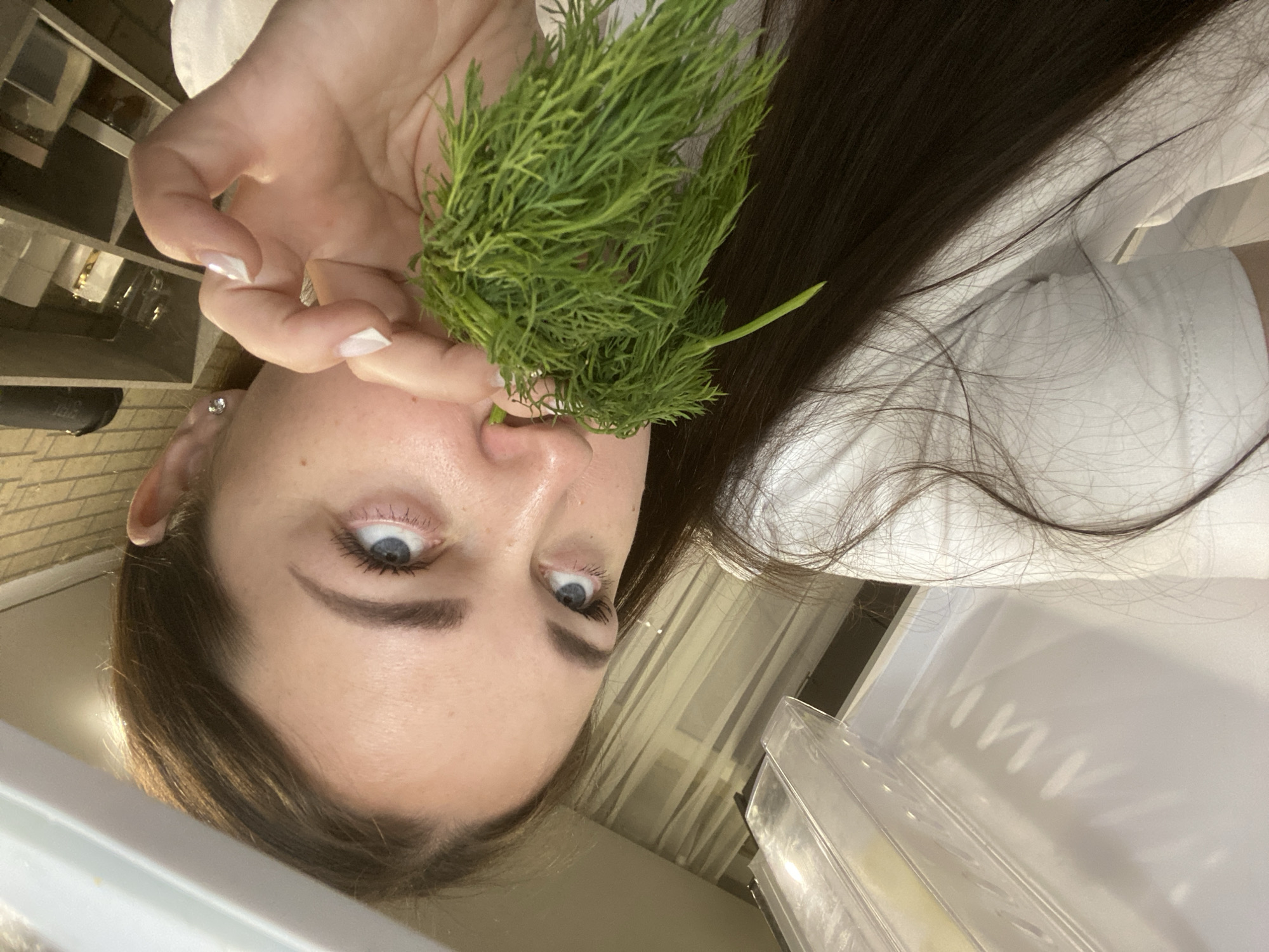 A woman holds a bunch of dill to her nose. She is wearing a white shirt and has long brown hair. The background is a kitchen setting, and there is a shelf in the upper left of the frame. The location is not discernible in the image.