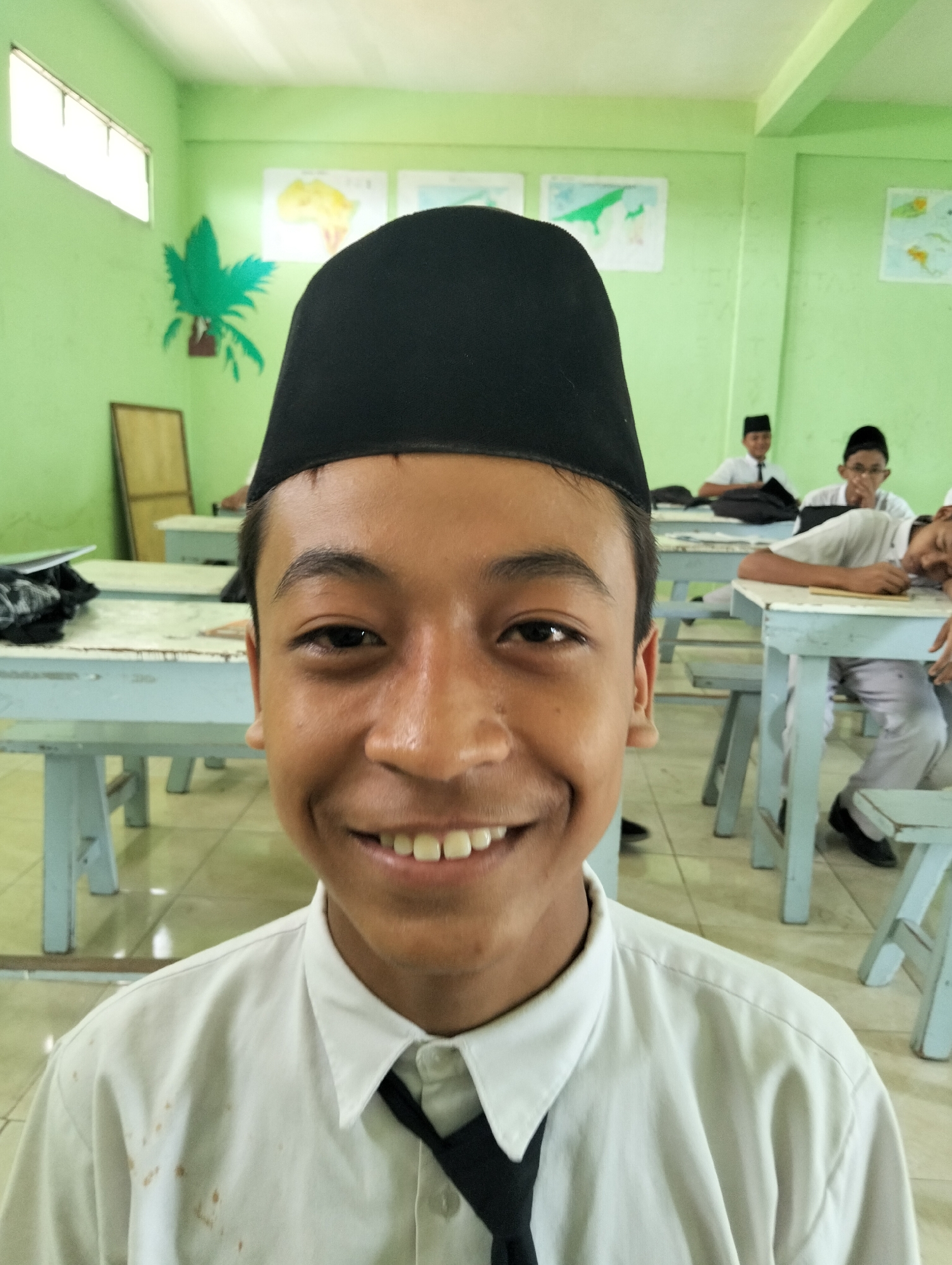 A young Indonesian boy smiles broadly at the camera in a brightly lit classroom. He is wearing a white collared shirt with a black tie and a black peci cap. The classroom walls are painted a pale green, with posters of animals and maps adorning them. Desks with light blue legs are arranged in rows, and other students can be seen in the background, some paying attention and others appearing to be dozing. The flooring is tiled and reflects the light. The scene appears to be within the city of Kranji, Indonesia.