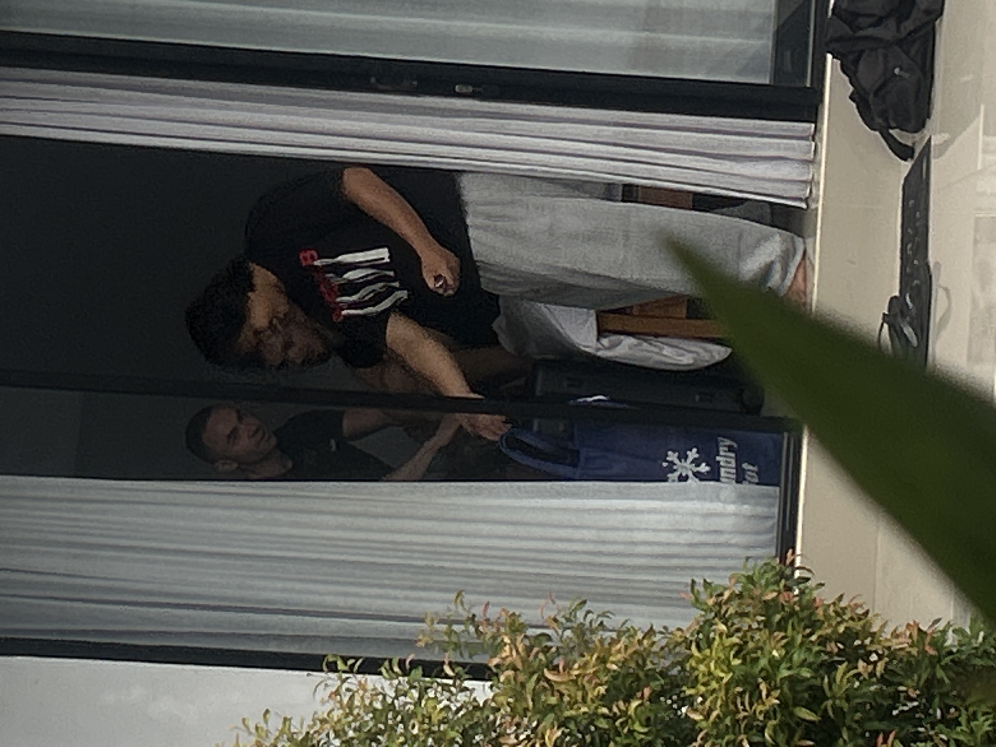In the city of North Kuta, Indonesia, at least three individuals are partially visible through an open doorway and adjacent windows. The scene appears to be outdoors, possibly a patio or balcony area, with white curtains to the left and right. In the center, a man wearing a black t-shirt and light grey sweatpants is interacting with a blue laundry bag that has white lettering and a snowflake design on it. To his left, another individual in a dark shirt is also engaged with the bag, with a portion of their arm visible. Further to the left and slightly behind them, a third person in a dark t-shirt is looking into the doorway. In the foreground, out of focus, is a lush green plant. On the tiled ground in front of the doorway, there are a pair of black flip-flops and a black backpack. The lighting suggests it is daytime.