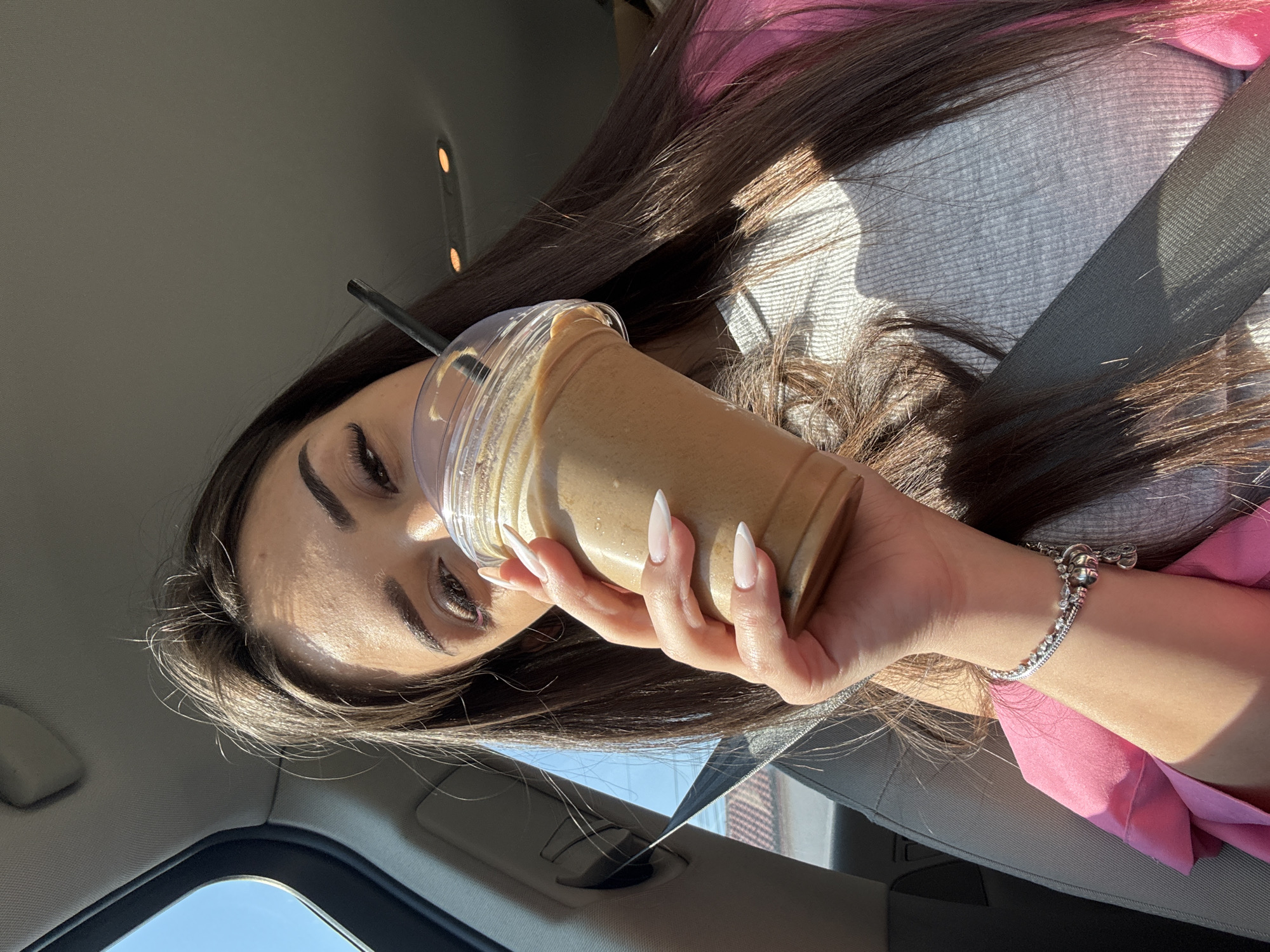A woman with long, dark brown hair is seated inside a vehicle, viewed from a slightly elevated angle. She is holding a clear plastic cup with a dome lid and a black straw, containing a light brown beverage, positioned in front of her lower face. Her left hand, adorned with a silver charm bracelet and featuring long, pointed nails with a French manicure, grasps the cup. Her eyes, framed by long dark lashes and defined eyebrows, are directed towards the viewer. A subtle pink eyeliner is visible on her lower eyelid. She wears a light grey ribbed top, with a bright pink garment draped over her left shoulder and arm. A dark car seatbelt crosses her chest. The car interior features a light grey fabric headliner with two amber overhead lights. Through an open window or sunroof on the left, a clear blue sky is visible, along with a partial view of a tiled reddish-brown roof and power lines in the distance. Strong sunlight illuminates the scene from the left, casting shadows. The event occurs in the city of Rakovski, Bulgaria.