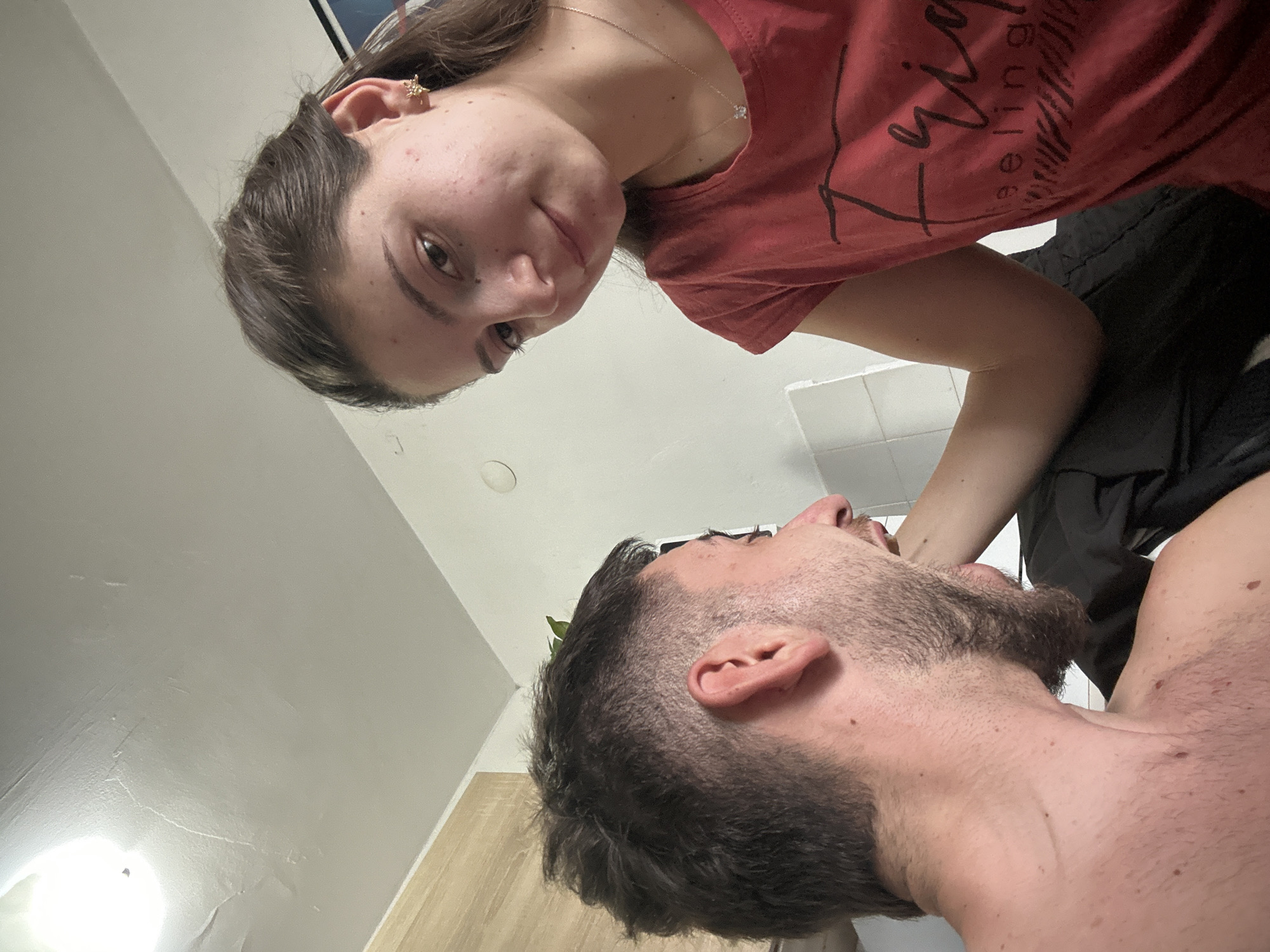 The image presents two individuals in what appears to be a bathroom setting. A woman with brown hair and wearing a pink t-shirt is positioned looking down at a man with a beard and short hair. He appears to be lying down with his head tilted upwards. The background features white tiled walls and a light-colored wooden floor, typical of a bathroom. The location is not identifiable as Sofia, Bulgaria based on the provided image.