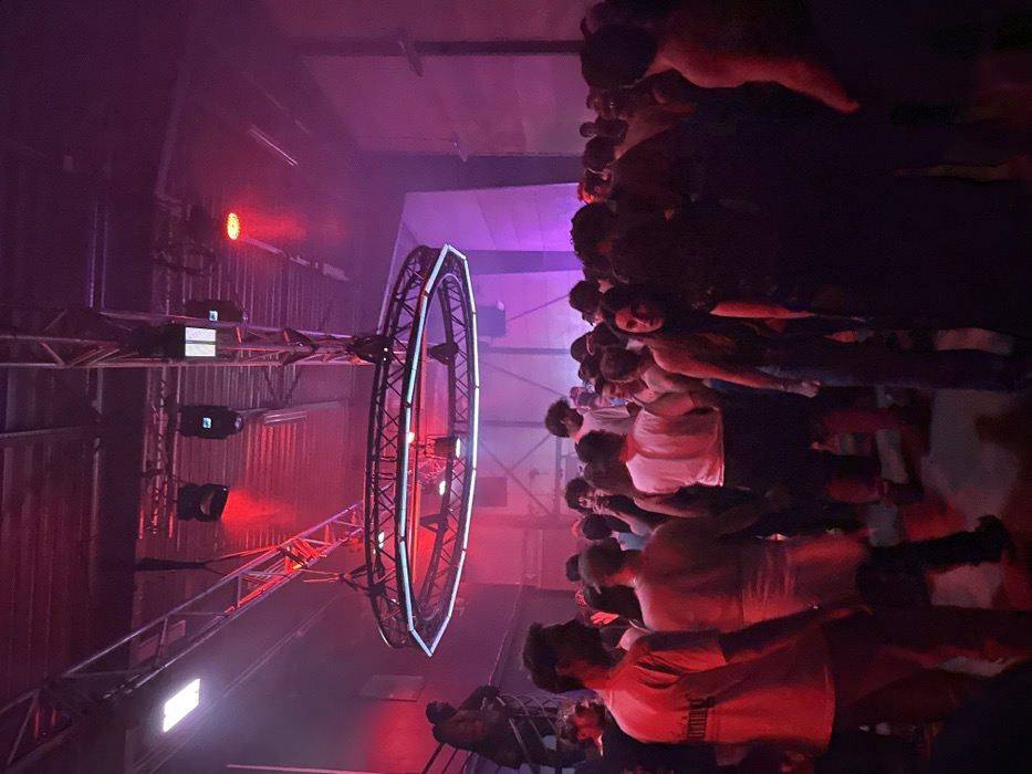 A crowd of people stands in a dimly lit space with a large, illuminated circular structure suspended overhead. The circular structure is attached to a metal truss system that also supports several lights. The lighting is primarily red and purple. Some attendees are looking in the direction of the camera. The overall setting evokes a sense of a concert, event, or club.