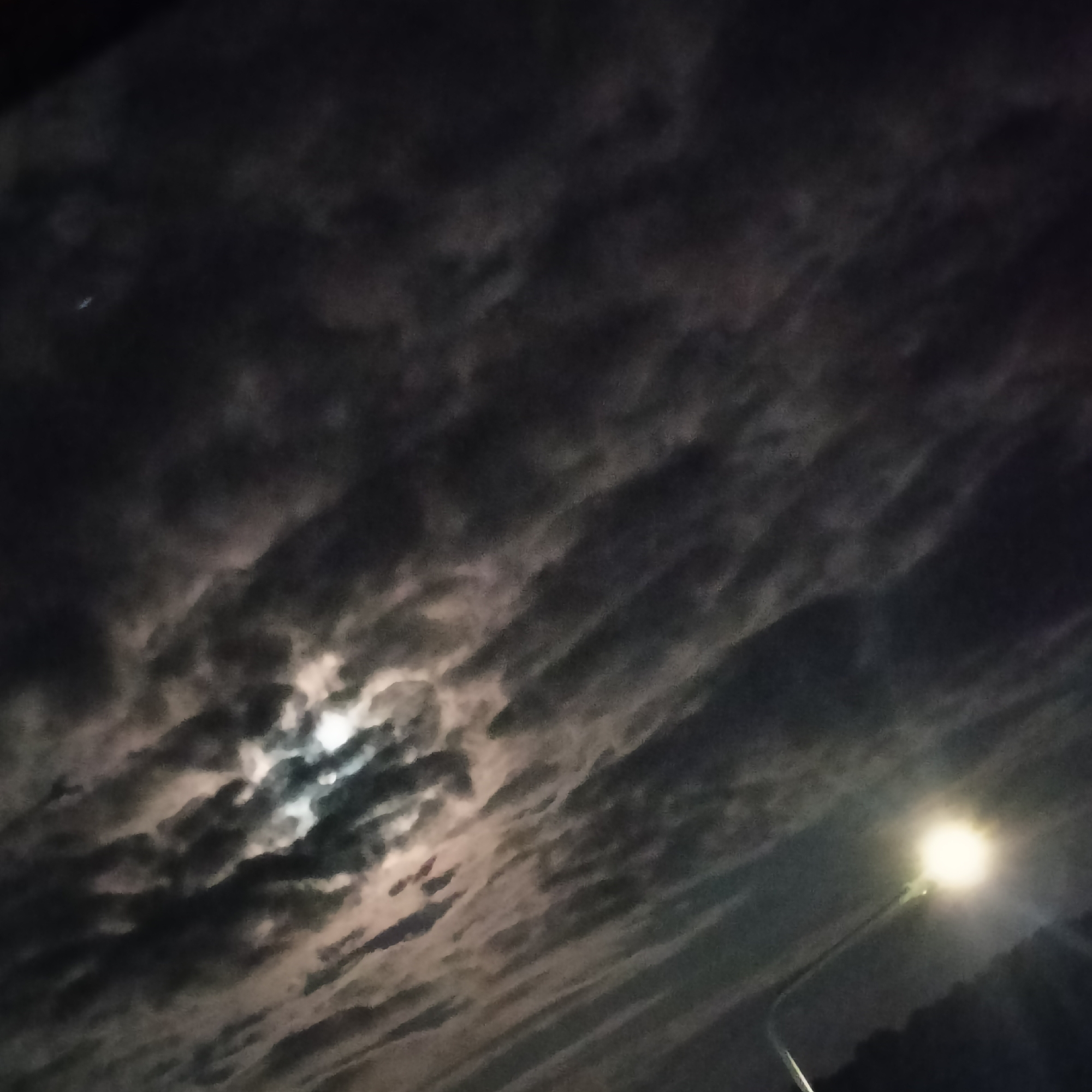The image captures a night sky over Limerick, Ireland, dominated by a scattering of dark, wispy clouds that partially obscure a bright, luminous moon. The moonlight filters through the clouds, creating a dramatic and textured sky. In the lower right quadrant of the image, a street lamp illuminates the scene with a bright, white glow, its pole extending upwards and out of frame. The overall impression is one of a cloudy night, with the moon and artificial light providing the primary sources of illumination.