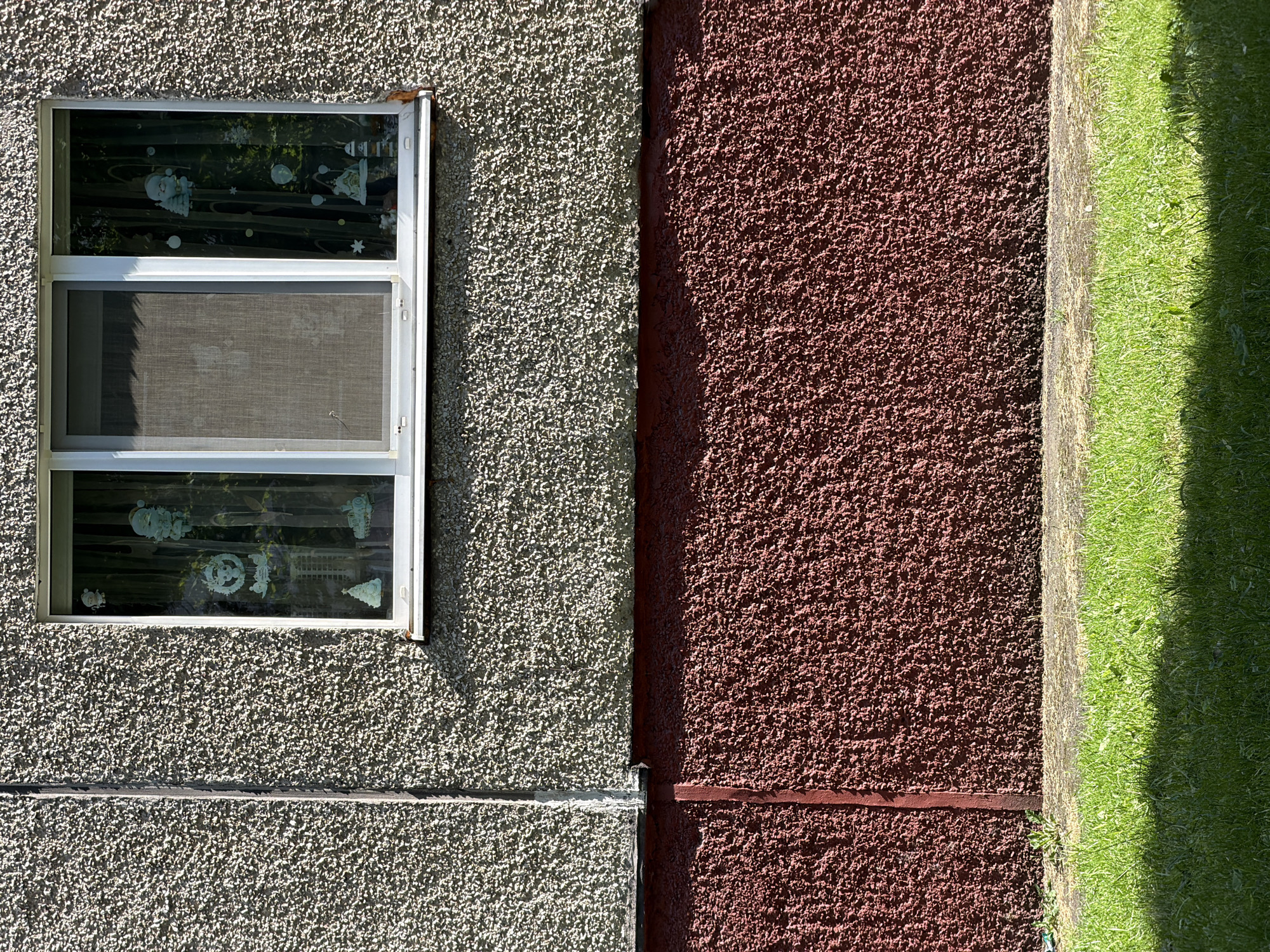 The image shows a section of a building exterior in Minsk, Belarus. The building has a rough, textured grey facade above a red, textured lower section. A sliding window with a white frame is visible in the upper part of the grey facade. Inside the right pane of the window, there are light-colored holiday decorations, including figures resembling Santa Claus, Christmas trees, and stars. The area below the building is paved, with a strip of green grass in the foreground, part of which is in shadow. The scene appears to be an outdoor residential area on a sunny day.