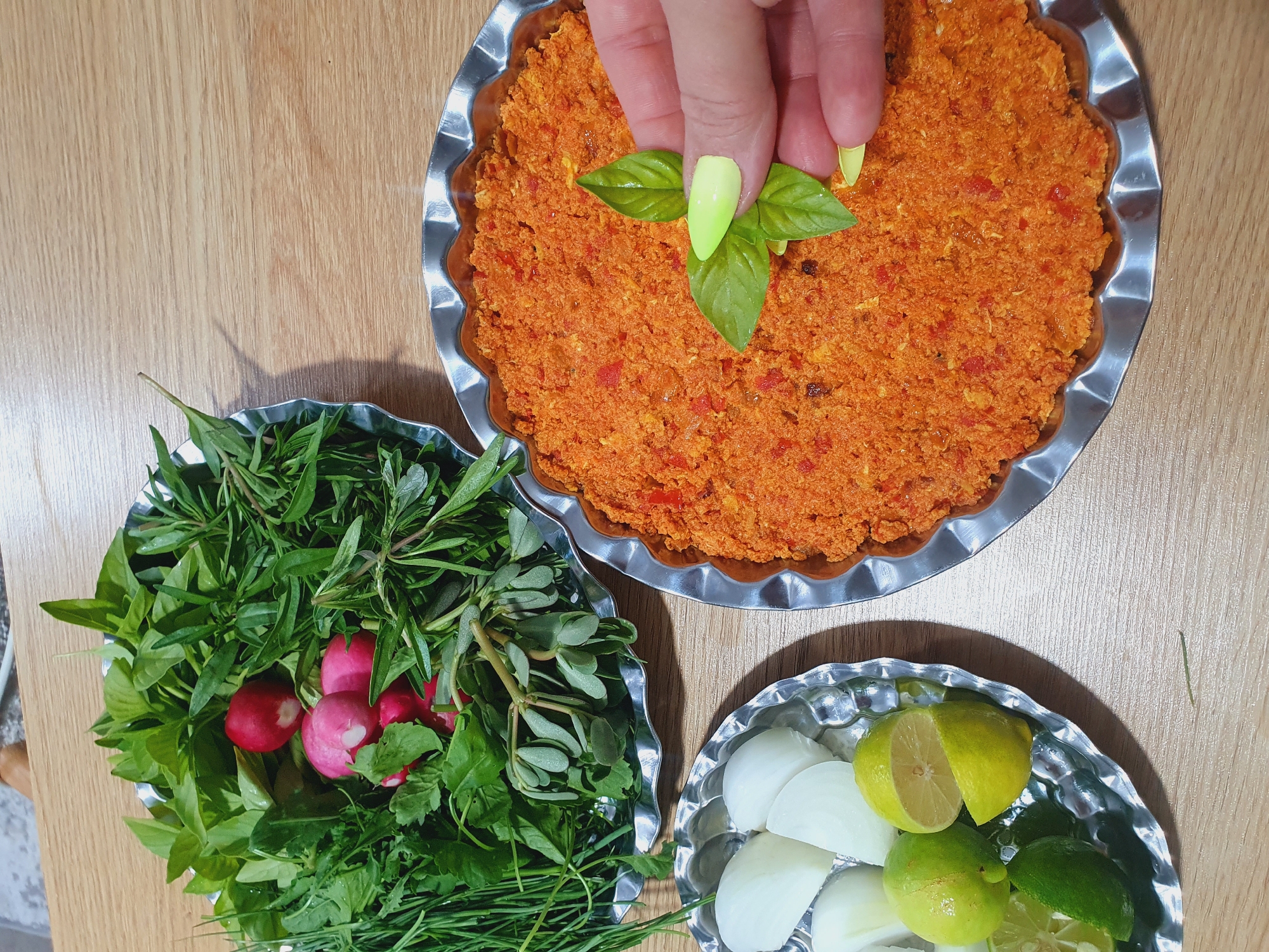 A high-angle, medium shot captures a table laden with food preparations on a wooden surface. In the top left, a fluted silver dish holds a vibrant arrangement of fresh greens and several radishes. To the right, another fluted silver dish contains a finely ground, orange-red mixture, possibly a spread or dip. A hand with bright yellow nail polish is shown delicately placing a fresh green basil leaf onto this mixture. In the bottom left, a third fluted silver dish presents chopped white onions and lime wedges. The scene appears to be a domestic setting, possibly in a kitchen or dining area.