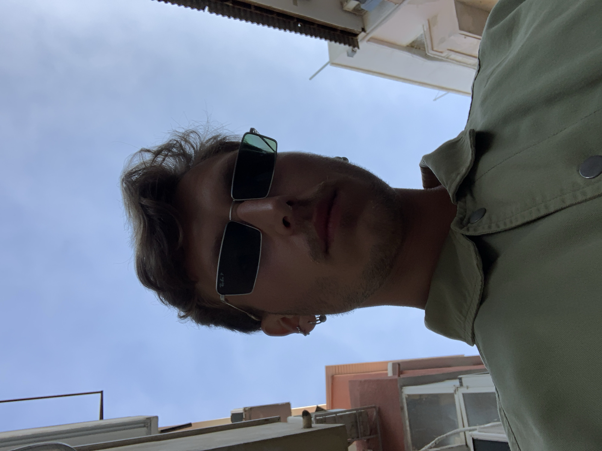 A low-angle shot shows a man wearing sunglasses and a light green shirt. He has brown hair and a short beard. In the background are buildings and a clear sky, possibly located in Tel Aviv, Israel.