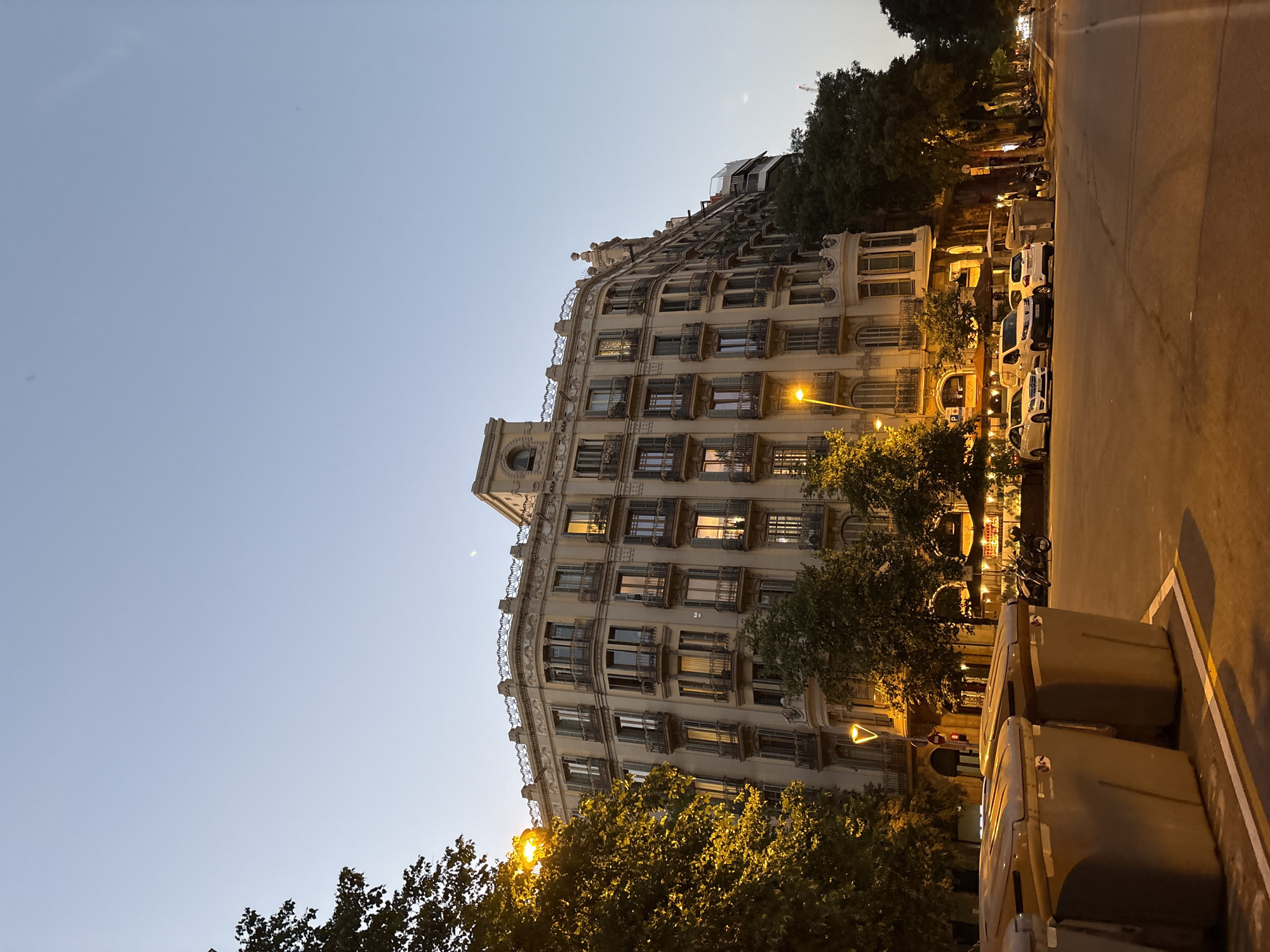 The image shows a street scene in Barcelona, Spain at dusk. A large, ornate building with multiple balconies and windows dominates the right side of the frame. The building appears to be residential or commercial, with lights on in some of the windows. Trees line the street in front of the building, and a street lamp illuminates the foliage with a warm yellow glow.
In the foreground, two large, grey trash bins are visible on the left side of the image. The street is paved, and several cars are parked along the side of the road. A motorcycle is also parked near the bins. The sky is a clear, pale blue, indicating the transition from day to night. The overall impression is one of urban life in Barcelona, with a blend of historic architecture and modern elements.