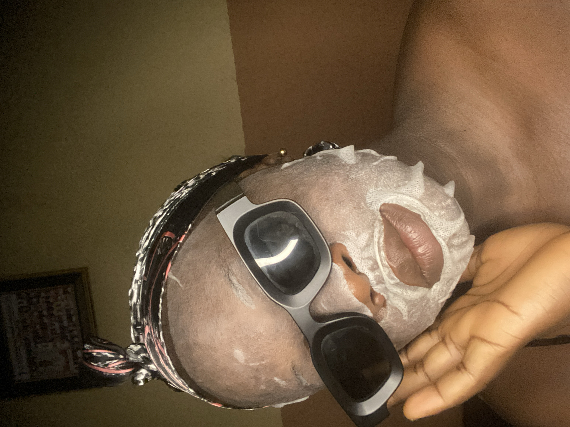 An image, rotated clockwise, captures a person of color engaged in a self-care routine in what appears to be an indoor setting in Maigizo, Nigeria. The subject's face is largely covered by a white sheet mask, which conforms to the contours of the skin, leaving an opening for the lips. Dark-lensed, grey-framed sunglasses or goggles are worn over the mask, covering the eyes. Braided hair is visible at the side of the head, secured by a patterned headwrap featuring black, white, and red/pink elements. A small gold-colored earring is discernible near the ear.
The person's right hand, with fingers slightly curled, rests gently near their jawline/cheek. The background is dimly lit, showing a plain wall in a muted, warm brown or olive-green hue. A portion of a framed picture, with faint details, hangs on the wall in the lower left corner. The overall ambiance suggests a private and relaxed moment, possibly in the evening or under low-light conditions, focused on a beauty or wellness treatment. There is no visible text in the image.