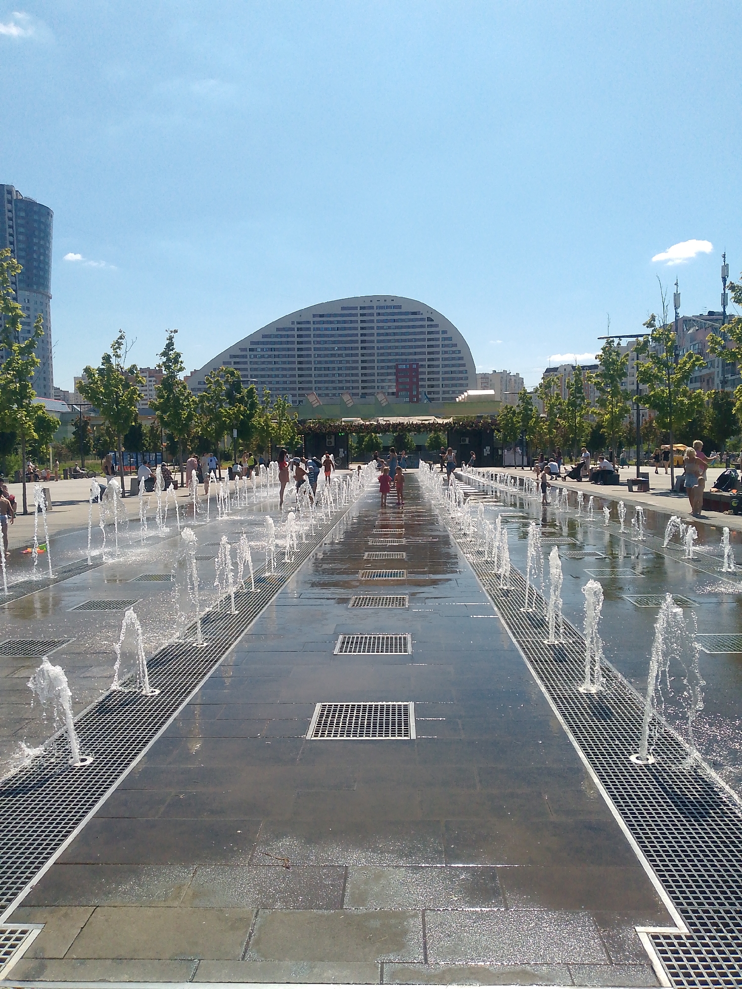 The image depicts a sunny day at a public plaza in Moscow, Russia, featuring a large, modern fountain with numerous jets of water spraying upwards from the ground. Several people, including children, are interacting with the fountain, some running through the water and others observing from the sides. The plaza is paved with large, dark tiles and incorporates metal grates for water drainage. In the background, there are trees, buildings, and what appears to be a large, curved, modern architectural structure. The sky is clear and blue.