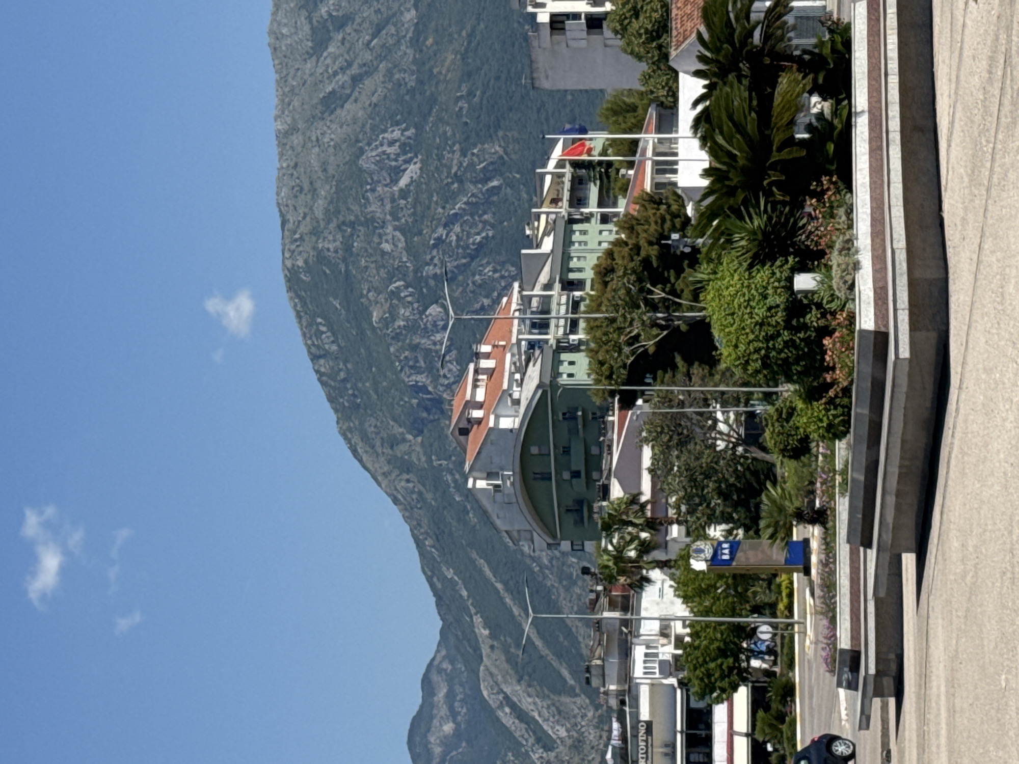 The image shows a street scene in the city of Bar, Montenegro. In the foreground, there are concrete benches and planters with greenery. A dark colored car is visible on the left. In the middle ground, there are several buildings, including one with a green facade and a red-tiled roof. Two flags are flying on poles in front of the buildings. A tall sign with the word "BAR" is also visible. In the background, a large, tree-covered mountain looms under a clear blue sky with a few scattered clouds. The overall impression is a sunny day in a coastal city.