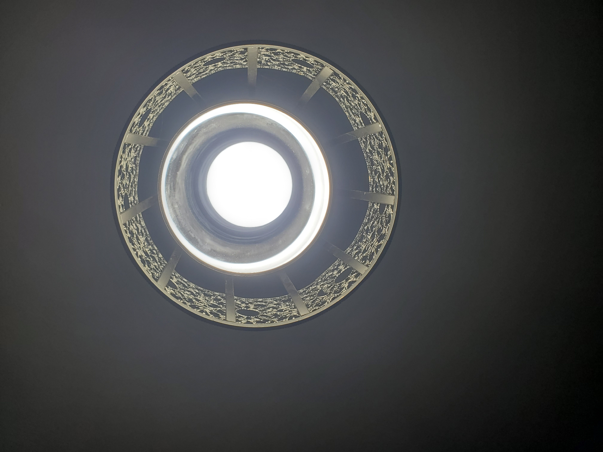 A circular, ornate light fixture is centered on a dark, textured ceiling. The fixture has a silver, filigree-like outer casing with a white, glowing light source visible in the center. The lighting creates a halo effect around the light and casts soft shadows on the textured ceiling.