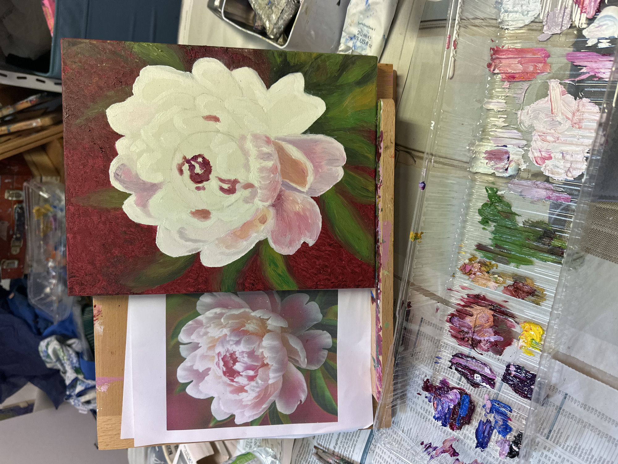 The image depicts an art studio setting, focusing on a painting in progress and the artist's tools. On an easel, a canvas displays a partially completed oil painting of a white peony against a dark red background. Beside the canvas, a printed reference image of a pink peony is placed on a wooden support. In the foreground, a clear plastic paint palette is filled with various shades of oil paints, including blues, purples, pinks, greens, yellows, and whites, suggesting the artist is actively working on the floral subject. The background is cluttered with art supplies, fabrics, and storage containers, indicative of a creative workspace. There is no discernible location within the city of Milan, Italy, or any specific geographical markers.