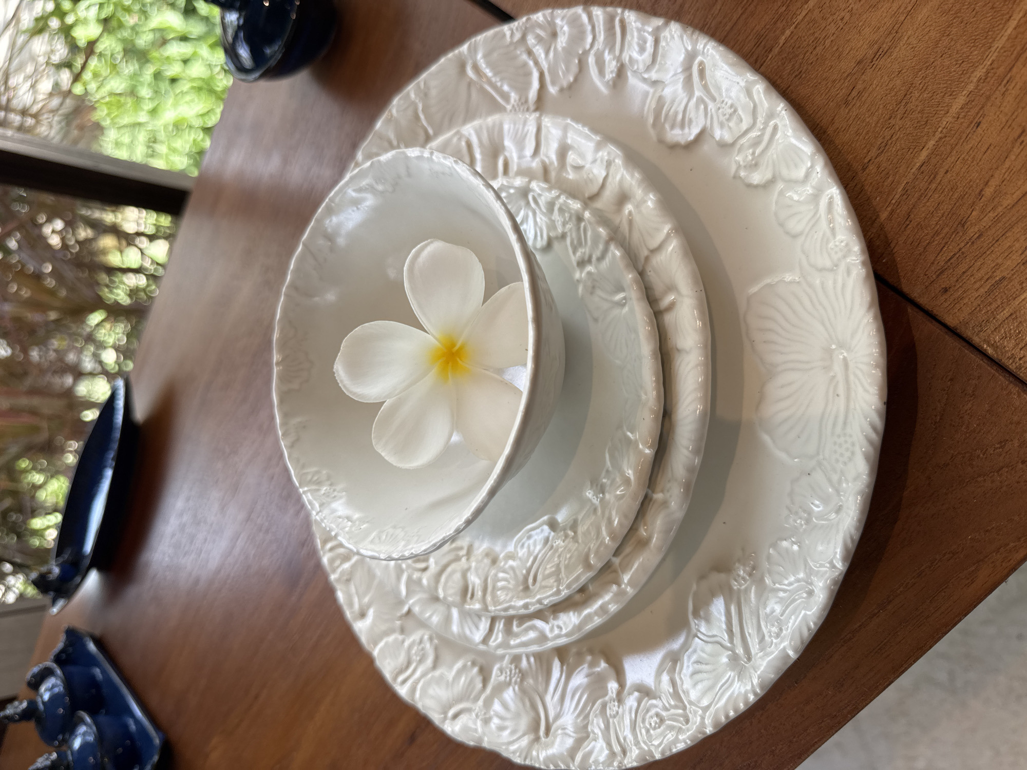 The image captures a table setting in Kuta Selatan, Indonesia. A stack of three embossed white plates sits on a wooden table. Atop the plates is a white bowl containing a single white and yellow plumeria flower. To the left of the plates are a partially visible dark blue decorative object and glimpses of an exterior scene, likely a window showing foliage. The scene is well-lit and appears to be indoors.