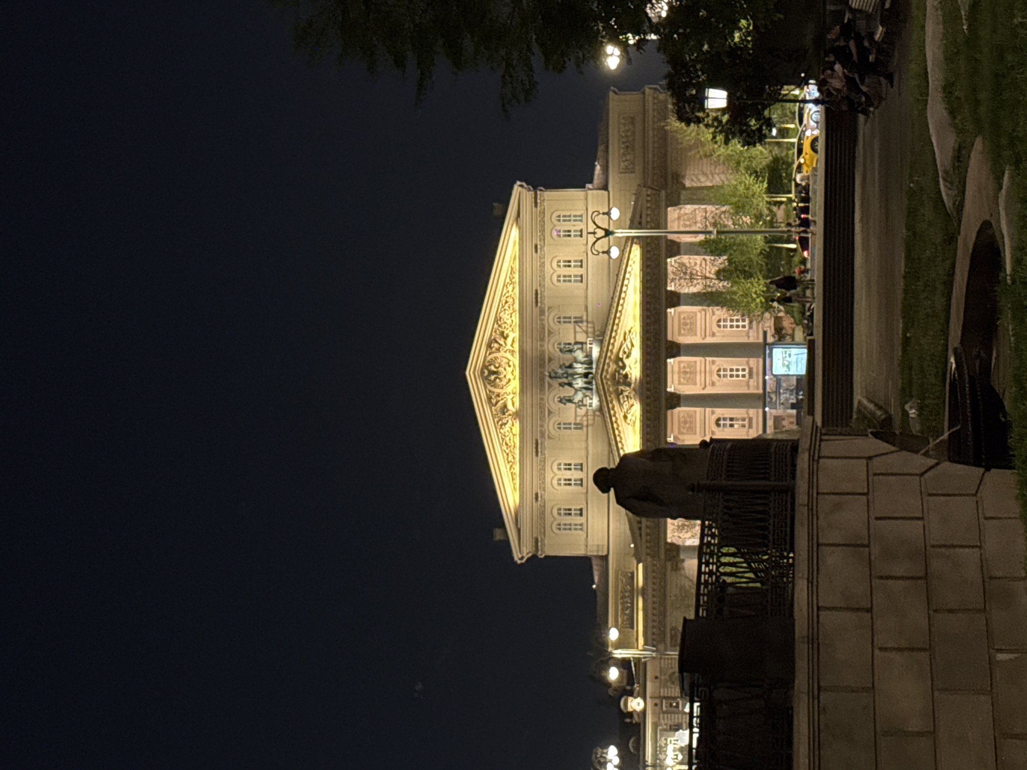 The image shows the Bolshoi Theater at night, located in Moscow, Russia. The building is illuminated with yellow light, highlighting its architectural details. A statue stands in the foreground. People are gathered on the steps near the theater. A few vehicles, one of them a yellow cab, are parked on the street behind the steps. Some trees and park elements surround the area. The sky is dark.