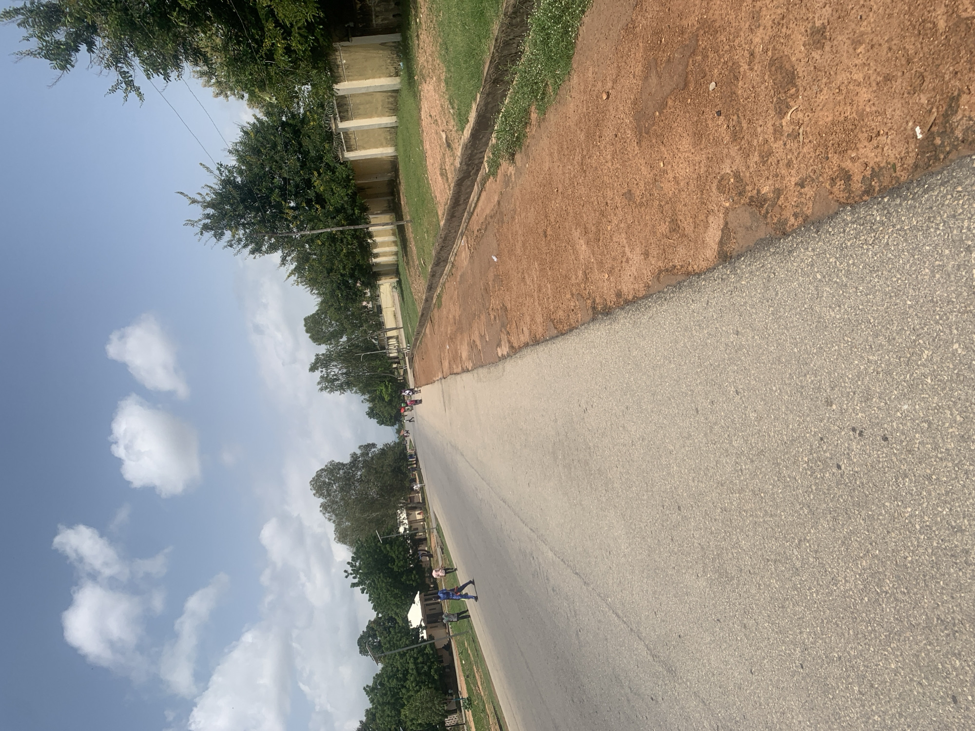 The image depicts a street scene in Jalingo, Nigeria, characterized by a paved road with a reddish-brown dirt shoulder. The road is lined with trees and some buildings on the left, while a wall with fence topping runs along the right side of the dirt shoulder. Several individuals are visible on the road and sidewalk, some walking and others standing. The sky is blue with scattered white clouds. The overall impression is one of a well-trafficked but not heavily congested urban area.