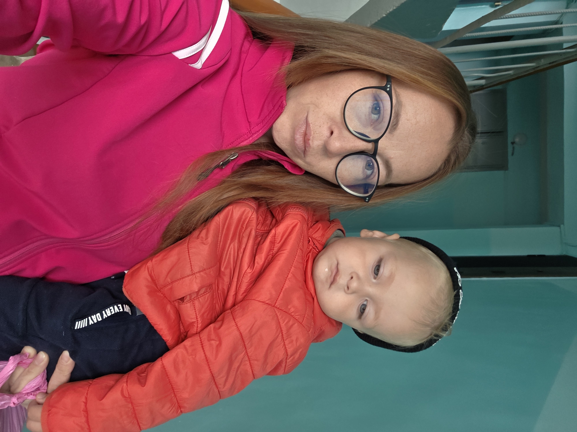 A woman with long, reddish-brown hair and glasses holds a young child. She is wearing a bright pink jacket. The child, wearing a black beanie and a red puffy jacket, is nestled in her arms. They are standing in what appears to be an indoor common area, possibly a stairwell or hallway, with a turquoise-painted wall in the background and a white railing and stairs visible to the left. The overall lighting is bright, suggesting it might be daytime.