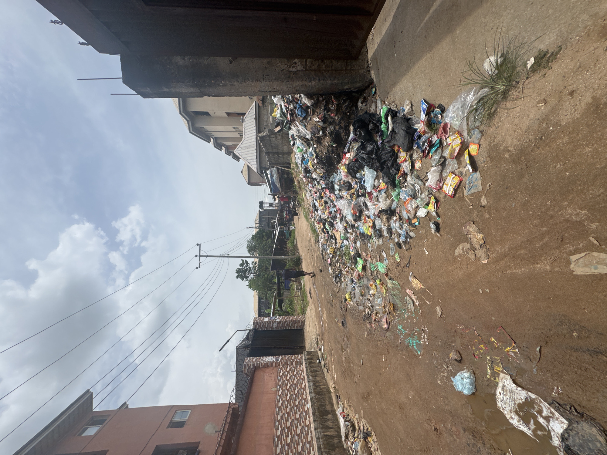 This image captures a wide outdoor scene, likely a street or alleyway, in Ihiagwa, Nigeria, characterized by a significant accumulation of trash.
The central subject is a very large pile of diverse refuse, dominated by countless plastic bags, food wrappers, and general household waste, heaped along the right side of a dirt path or road. More scattered trash, including colorful plastic pieces and wrappers, lies on the muddy ground, alongside small puddles and patches of green weeds.
On the left side of the frame, there are several buildings, some appearing residential. One building has a reddish-brown facade, while another is lighter-colored. A prominent feature is a sturdy-looking dark metal gate, topped with barbed wire, set into a patterned brick or tiled wall. Further down the path, a single person, dressed in dark clothing, is seen walking away from the viewer, near a wooden power pole. Multiple overhead power lines stretch across the sky from this and other unseen poles.
The scene takes place during the daytime, indicated by the bright, partly cloudy sky. The ground is largely unpaved dirt, appearing muddy in areas. The overall impression conveyed is one of poor waste management and environmental neglect. No discernible text is clearly visible on any of the packaging or other objects.