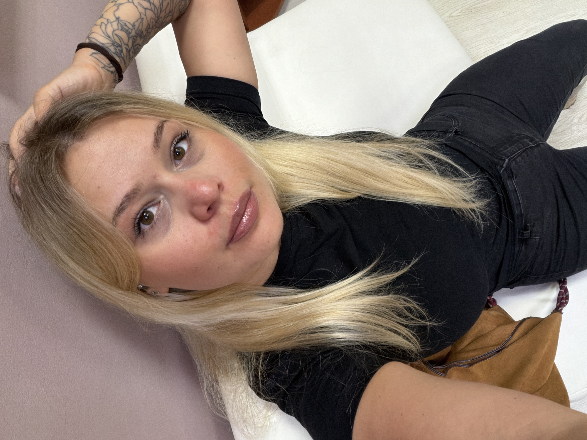 A woman with blonde hair is lying down, wearing a black top and black jeans. Her right arm, which features a tattoo, is raised, and her left hand is touching her hair. A brown bag is visible near her. The location is unknown.