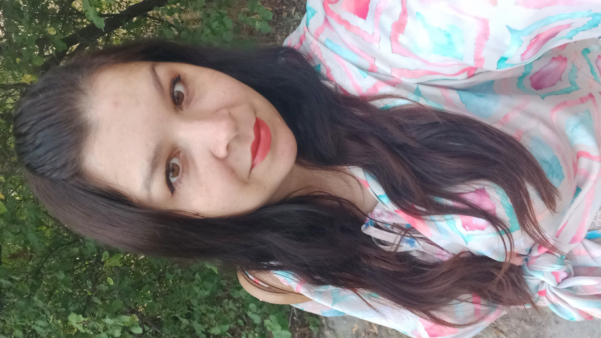 A woman with long, dark hair and brown eyes, wearing a colorful, patterned shirt, takes a selfie outdoors. She has red lipstick on. Behind her, there are green leaves and trees, suggesting a natural, outdoor environment. The overall scene appears casual and relaxed.