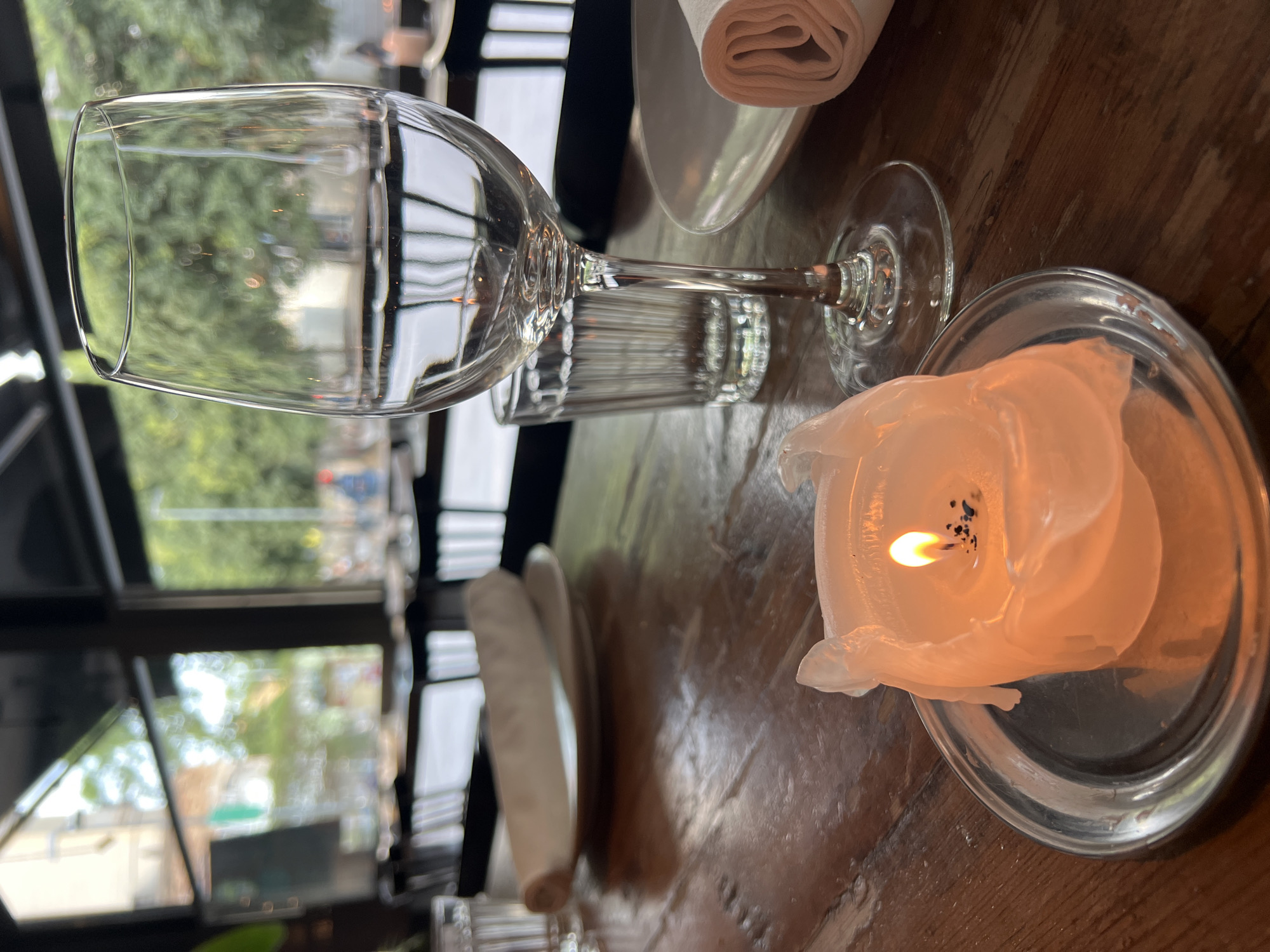A close-up shot shows a lit candle on a silver saucer on a wooden table in a restaurant in Barcelona, Spain. The candle is partially melted, with a flame burning brightly. A wine glass filled with water is also on the table, along with a smaller glass. In the background, there are other tables and chairs, and a window looking out onto a street scene with trees and buildings. The lighting is dim, suggesting it might be evening.
