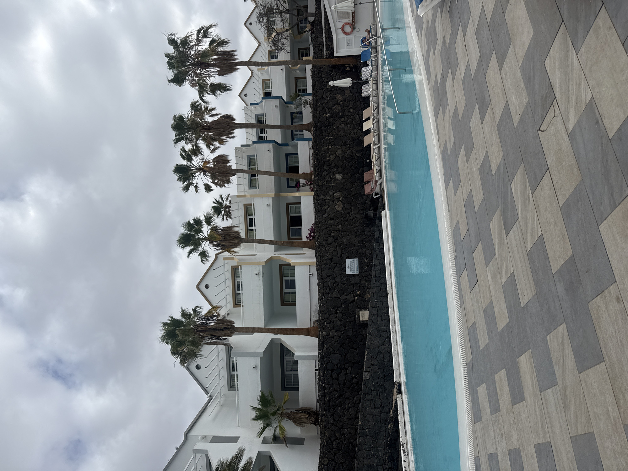 The image depicts a hotel or resort in Puerto Del Carmen, Spain, recognizable by its white, multi-story buildings with gabled roofs and blue trim, commonly found in this location. Palm trees are interspersed between the buildings and a black lava rock wall. Adjacent to the wall is a swimming pool with light blue water, alongside a paved area with a geometric pattern of grey tiles. The sky is overcast. No people are immediately visible in the frame. The scene suggests a calm and possibly off-season atmosphere at the resort.