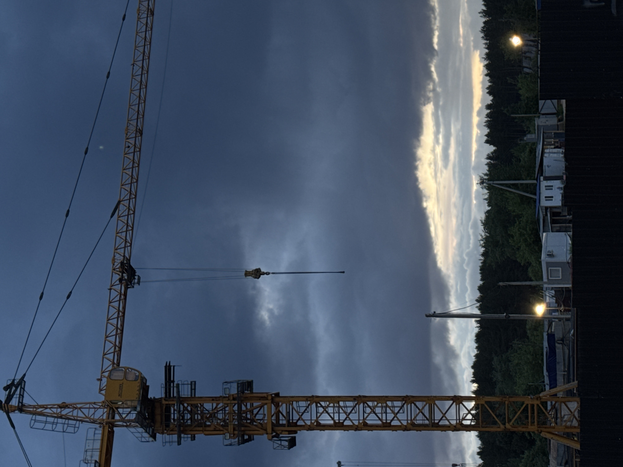 The image shows a yellow tower crane against a dark, cloudy sky. A portion of forest, buildings, and lights are visible in the background on the right side. The location is Izhevsk, Russia.