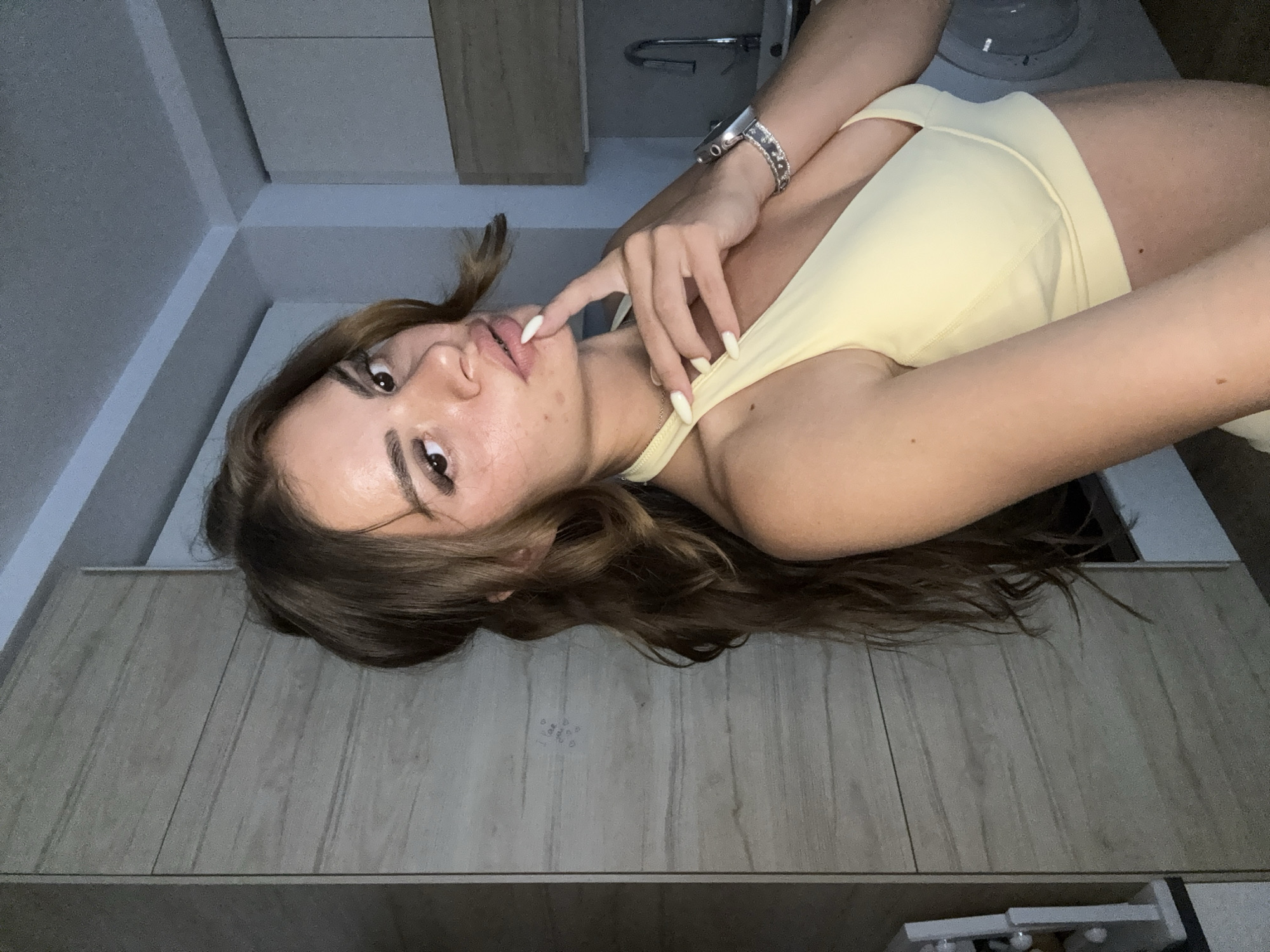 The media file shows a young woman lying on a wooden surface. She is wearing a light yellow crop top. Her hair is long and brown, falling around her shoulders. Her left hand is near her face, with her fingers touching her lips. She is looking towards the camera with her eyes open. She is wearing makeup, including mascara and eyeliner, and her eyebrows are well-defined. Her nails are painted white. She is also wearing a watch on her left wrist. The background appears to be indoors, with some cabinetry and a sink visible. The lighting suggests it could be daytime. The image has a slightly angled perspective, as if taken from above. The setting appears to be a domestic environment, possibly a bathroom or kitchen. No visible text is present in the image. The overall emotion conveyed is one of casualness and self-awareness.
