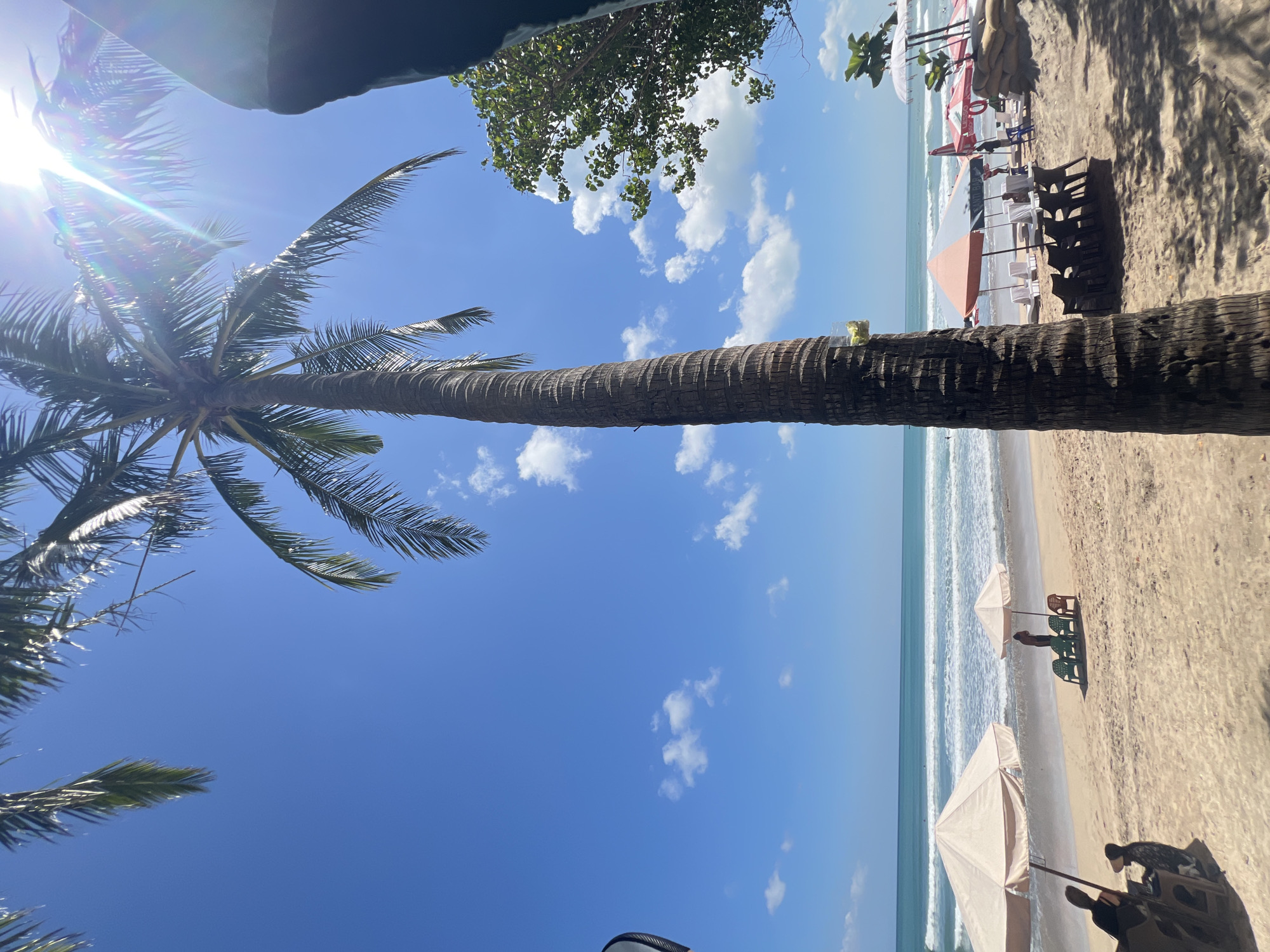 The image depicts a bright, sunny day on Kuta Beach, Indonesia. A tall, textured palm tree trunk dominates the foreground, casting dappled shadows on the sandy beach. The ocean stretches out to the horizon with gentle waves rolling onto the shore. Several beach umbrellas, some light beige and others with orange and white patterns, are set up on the sand, providing shade. In front of some umbrellas, there are rows of empty dark plastic chairs. A few people are visible on the beach; some are seated under the umbrellas, while others are standing or walking near the water. The sky is a vivid blue with scattered white cumulus clouds. The sun is high in the sky, creating a lens flare effect in the upper right portion of the image.