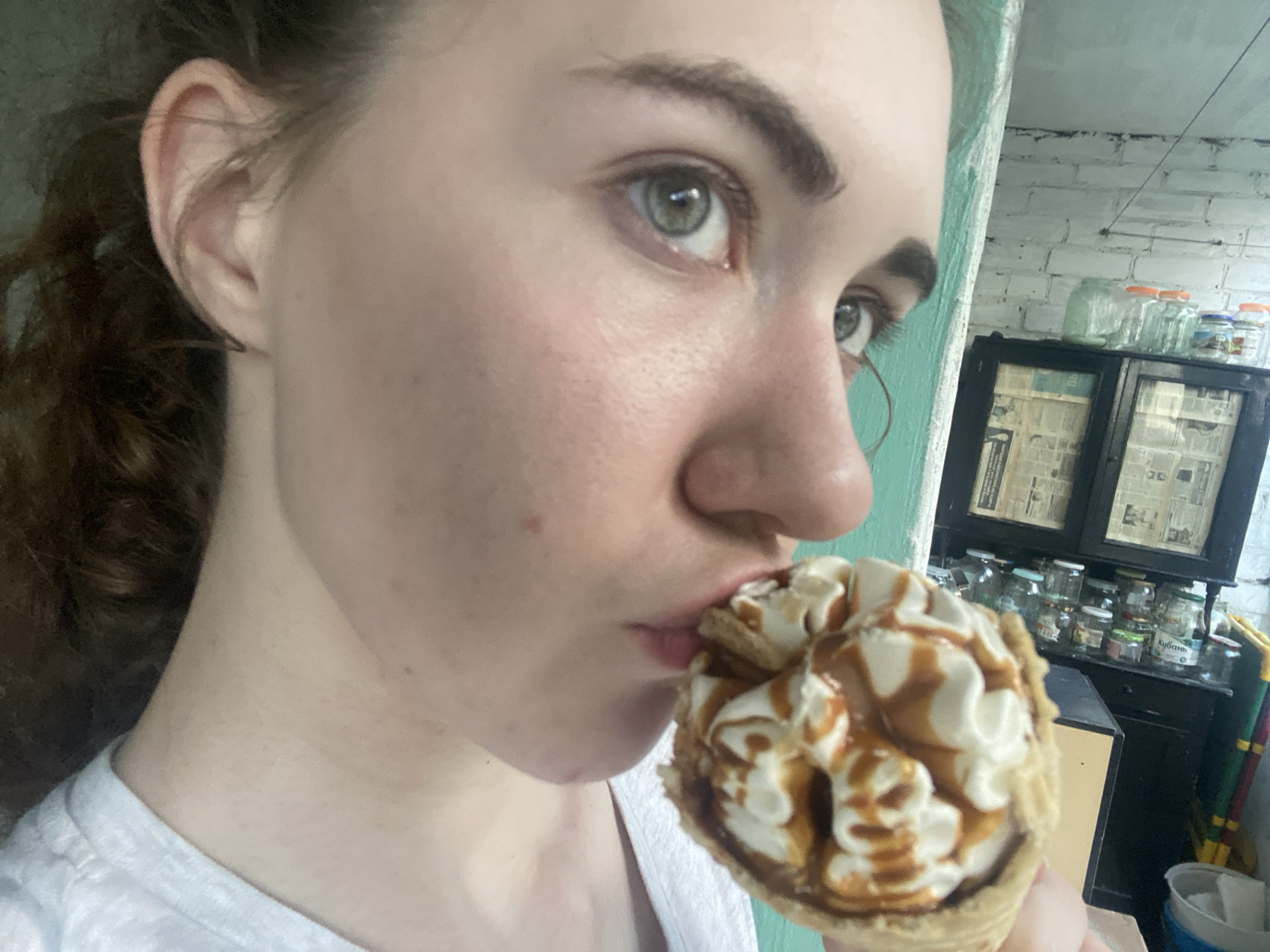 A close-up, profile shot shows a young woman with light skin, wavy brown hair pulled back, and green eyes as she eats an ice cream cone. She is wearing a white t-shirt. The background is out of focus, but appears to be a rustic interior with a pale green painted wall, a wooden shelf holding jars and what looks like a newspaper-lined cabinet. The overall scene is casual and appears to be indoors, possibly in a kitchen or pantry area. The photo is taken at an angle that emphasizes the woman's face and the ice cream.