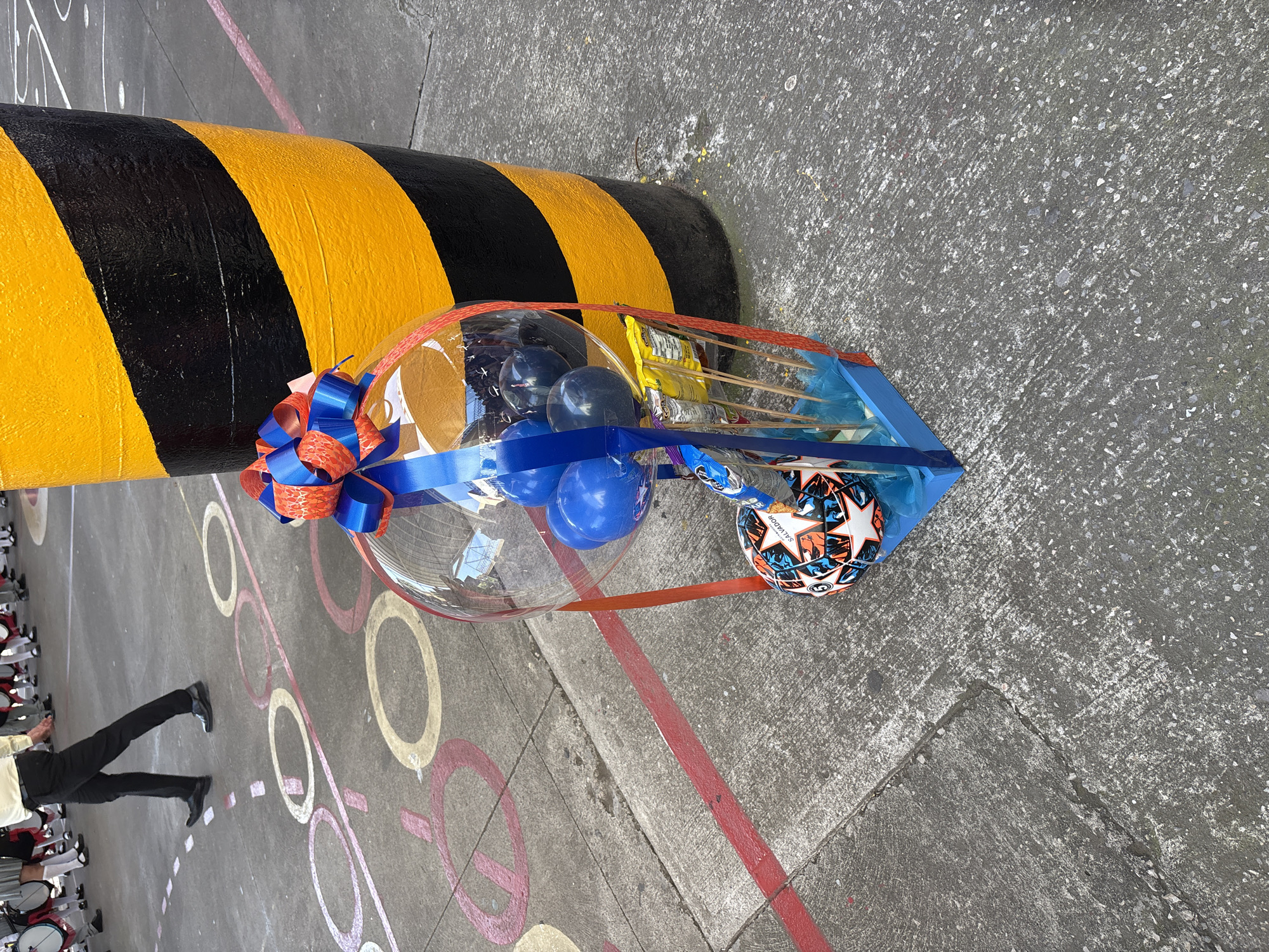 The image depicts a ground-level view of a courtyard or paved area, likely outdoors, with markings on the ground suggesting a sports court or a children's play area. The ground is concrete with various painted circles and lines in red, pink, and white.
In the foreground, to the right, is a gift basket presented in a clear plastic bubble. The basket is decorated with blue and orange ribbons. Inside the bubble, there are several blue and dark-colored balloons. At the bottom of the basket, a small soccer ball with a white, black, orange, and blue design is visible, along with several packets of snacks attached to wooden skewers. The basket is supported by a blue wooden base.
Behind the gift basket, a large, cylindrical object is prominently featured. This object is painted with thick diagonal stripes of bright yellow and black, resembling a caution pole or marker.
To the left of the gift basket and pole, a person, likely a man, is walking away from the camera. They are wearing black pants, a light-colored collared shirt, and black dress shoes. Their posture indicates movement, and their attire suggests they might be part of an organized group or event.
In the background, on the left side of the image, a group of people can be seen. They appear to be wearing uniforms, including white shirts and dark pants or skirts, and some are holding large drums. This suggests a marching band or a similar musical ensemble. The overall setting implies a public event or gathering. The style of the painted markings on the ground and the presence of a marching band could be indicative of a school or community event in Tejalpa, Mexico, although this cannot be definitively confirmed by the image alone.