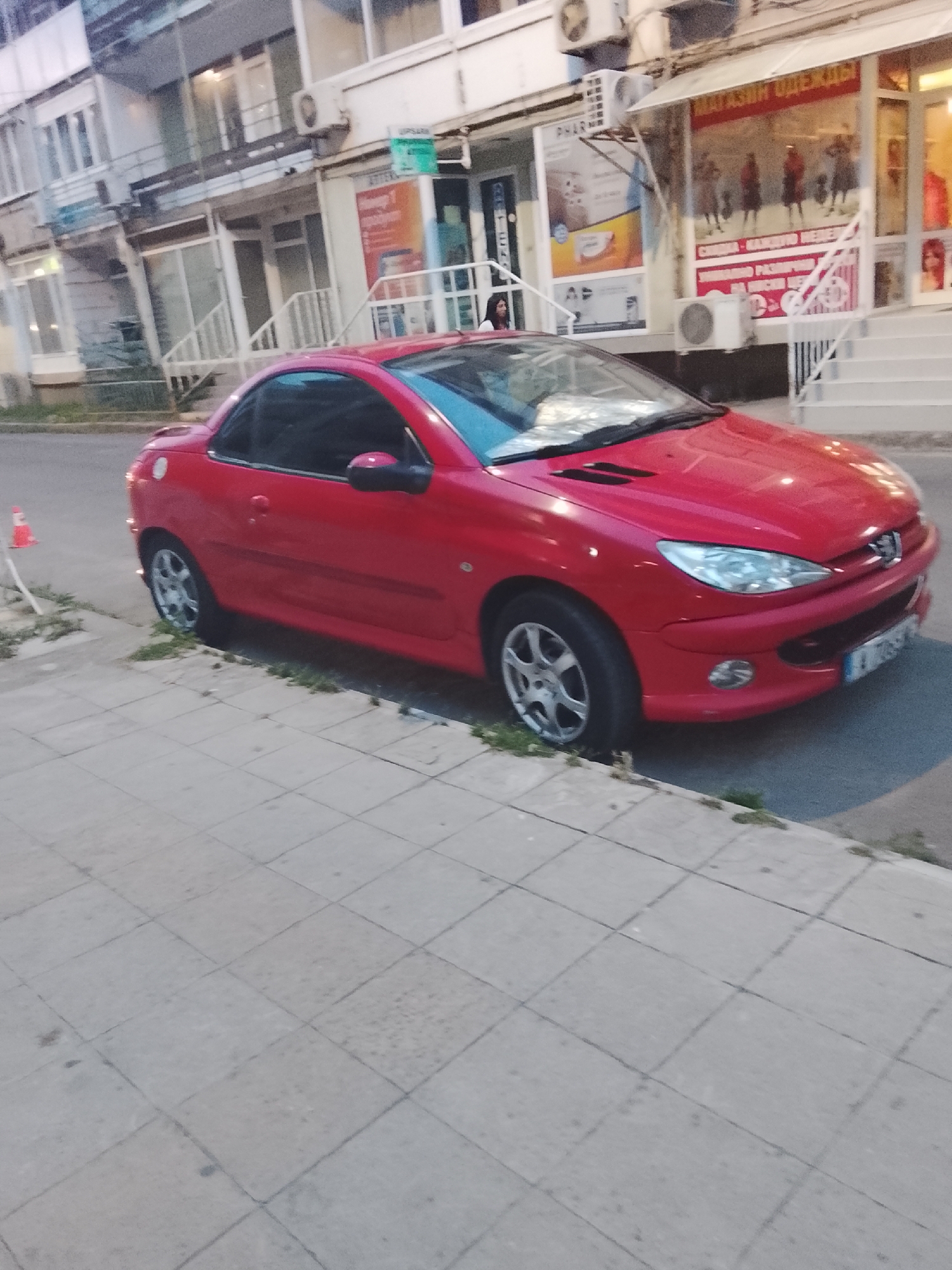 A red Peugeot 206 CC convertible is parked on a sidewalk next to a street in Nesebar, Bulgaria. The car is positioned at an angle, facing towards the right of the frame. The sidewalk is paved with grey square tiles. To the left of the car, a small orange traffic cone is visible near the edge of the sidewalk. Behind the car, a building with shops on the ground floor is present. One shop has a sign with Cyrillic text, and the windows display mannequins dressed in clothing. Another shop has a sign that reads "PHAR." An air conditioning unit is mounted on the exterior wall of the building. A woman with dark hair is standing near the entrance of one of the shops. The scene appears to be a commercial street in the city.