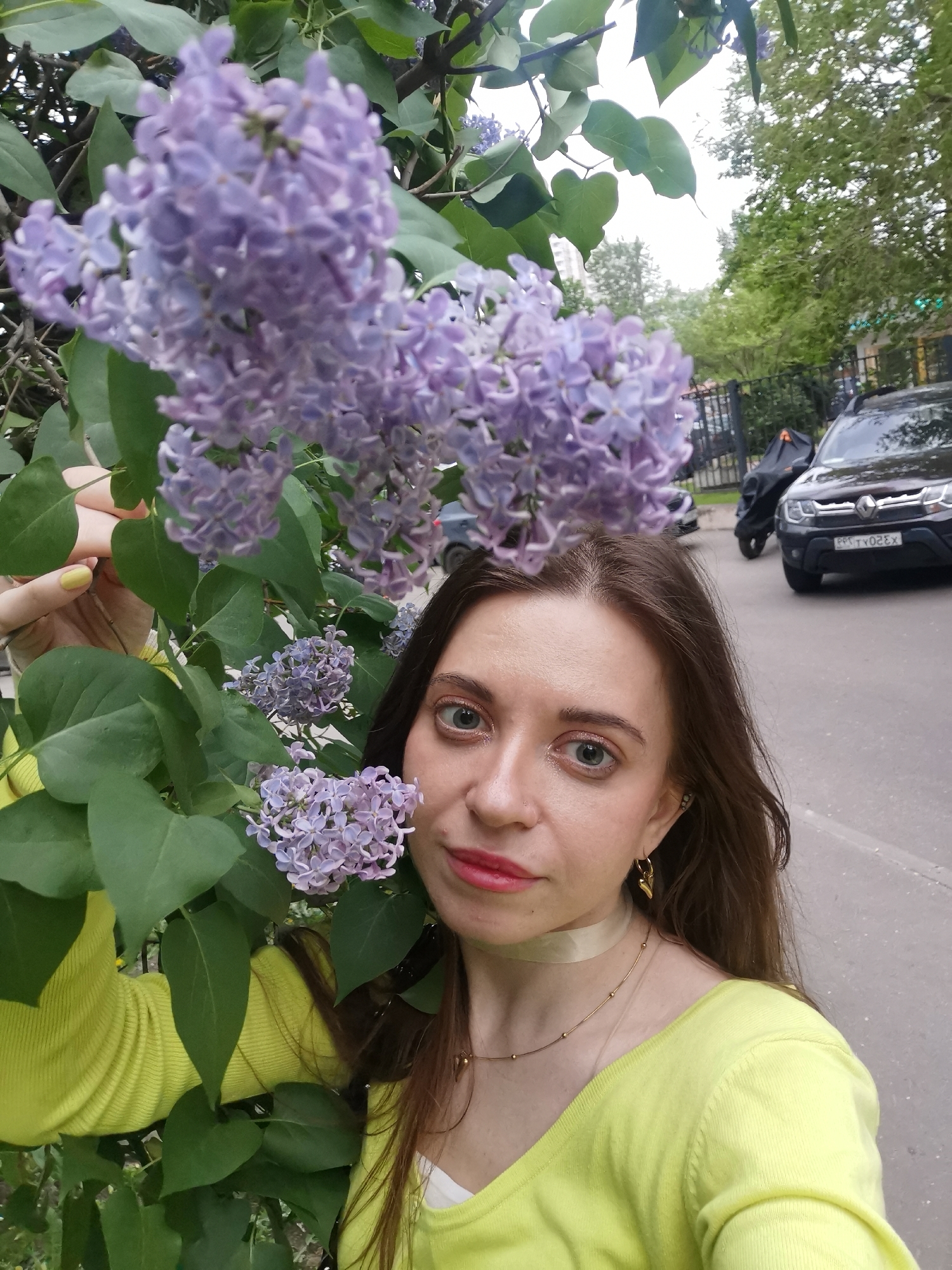 A woman is taking a selfie with lilac flowers. She is wearing a yellow sweater and has brown hair. There is a black SUV parked on the street behind her. The location appears to be a city street with trees and a fence. The city and country are Unknown.