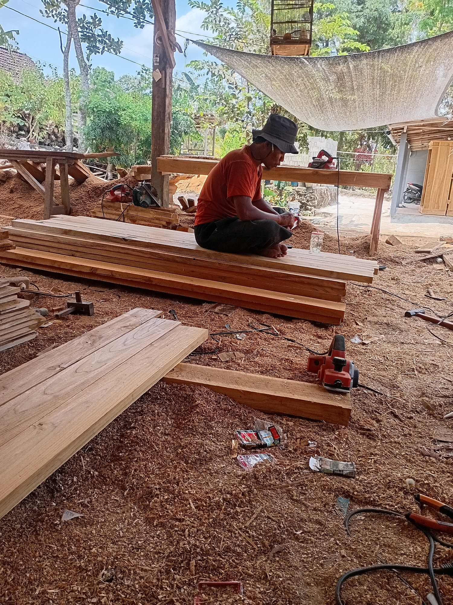 A man in a wide-brimmed hat and an orange shirt sits cross-legged on a pile of lumber, working on a piece of wood. He is surrounded by sawdust and woodworking tools, including a planer, saws, and hammers. In the background, stacks of lumber and a workbench are visible. The scene appears to be an outdoor workshop, likely in Playen, Indonesia, given the vegetation and the style of construction. A birdcage hangs from a wooden beam overhead.
