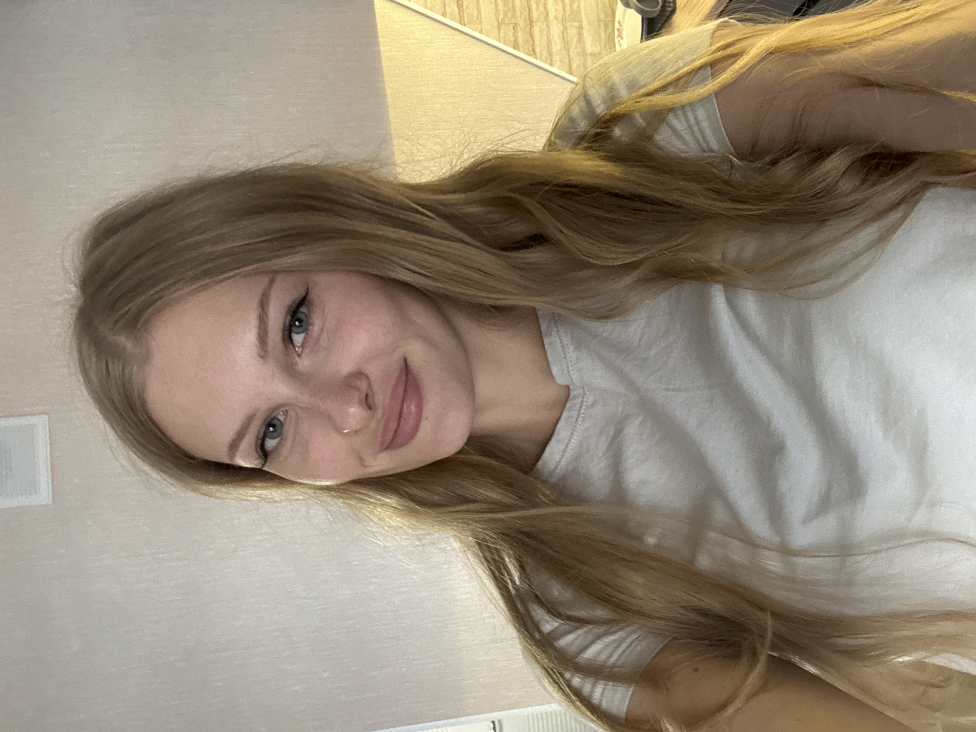 A close-up, head-and-shoulders selfie of a young Caucasian woman with long, wavy blonde hair and blue eyes. She is wearing a light gray t-shirt. She has light makeup on, including eyeliner and lipstick, and a small nose ring. She is smiling faintly. The background is a textured, light-colored wall. To the right, there is a bright yellow, angled surface, possibly a wall or partition, with a textured pattern. The overall lighting suggests an indoor setting, likely residential. There are no discernible landmarks or features that would specifically place the scene within the city of Perm, Russia.