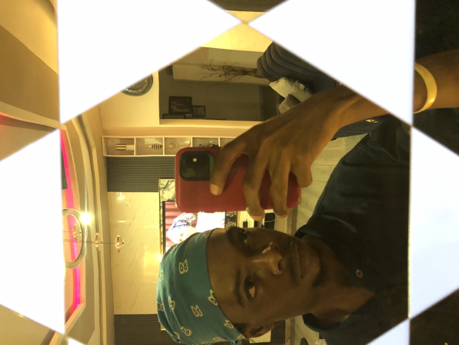 This image captures a close-up selfie reflection of a male individual, presumably located within an indoor residence in Tema, Ghana.
**Subject:** A dark-skinned male is depicted from the chest up, with a direct but slightly averted gaze to his left. He is wearing a light blue bandana with white patterns on his head and a dark blue collared polo shirt. A yellow wristband is visible on his right wrist, which is holding a red-cased smartphone. His left hand is not visible.
**Action:** The male is actively taking a selfie photograph using the red-cased smartphone, which is held by his right hand and positioned in front of his face. The phone's camera lens is visible.
**Environment (reflected in the mirror):** The selfie is taken in a mirror composed of white triangular segments, creating a fragmented view of the surroundings.
* **Ceiling:** A modern ceiling features a central circular light fixture, possibly with multiple suspended pendant lights, and ambient pink or magenta strip lighting recessed into a false ceiling structure.
* **Walls:** The background wall behind the subject displays a section with vertical dark gray paneling, adjacent to lighter-colored walls.
* **Television:** A large flat-screen television is mounted on the wall, displaying an image of a person, possibly a woman, also wearing a head covering.
* **Shelving:** To the right of the TV, a tall, narrow white shelving unit contains several decorative objects, including what appears to be metallic vases or statues.
* **Further Room:** Beyond the shelving unit, another room or hallway is visible, featuring a dark wall clock, framed pictures, and a light-colored couch with multiple throw pillows. Decorative dried branches are visible near the couch.
* **Lighting:** The scene is illuminated by warm, artificial interior lighting.