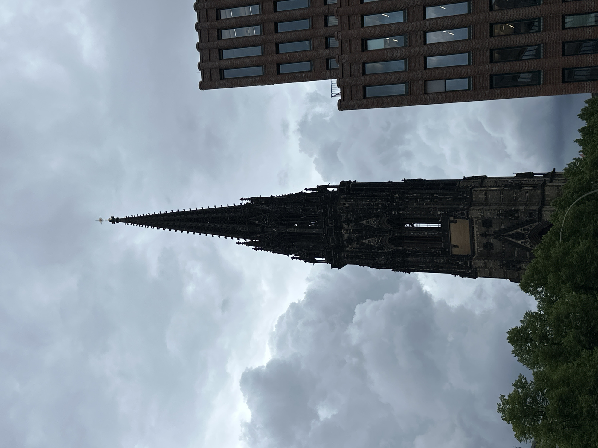 The image captures an urban architectural scene in Hamburg, Germany, under a dramatic, overcast sky. A towering, slender Gothic church spire, dark in color and intricately detailed with ornate stonework, dominates the left side of the frame, reaching upwards towards the clouds. A small cross is visible at its pinnacle.
Contrasting sharply with the ancient church, a modern brick building occupies the upper right portion of the image. This structure features a textured facade of dark reddish-brown bricks, punctuated by numerous rectangular windows that reflect the grey light of the sky. The design appears functional and contemporary, providing a stark architectural juxtaposition with the historic spire.
The sky is a prominent element, filled with heavy, swirling grey clouds that create a moody and atmospheric backdrop. The overcast conditions suggest a cloudy daytime, possibly hinting at an impending storm or simply a generally grey day. In the bottom right corner, a section of vibrant green foliage from a tree is visible, adding a touch of natural color to the otherwise dark and muted palette of the buildings and sky. No people or discernible activities are present, and there is no visible text. The scene emphasizes the unique blend of historical and modern architecture common in European cities.