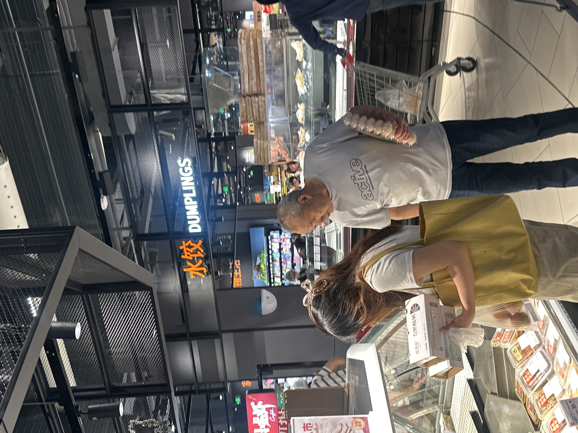 In a supermarket in Chongqing High-tech Zone, China, an elderly man wearing a white t-shirt with the word "active" printed on it is holding a package of dumplings. He is standing next to a shopping cart. In front of him, a woman with long brown hair is holding two boxes and a plastic bag. She is wearing a white t-shirt and a yellow tote bag. Behind them, a sign reads "水饺 DUMPLINGS". The supermarket appears to be well-lit and stocked with various food items.
