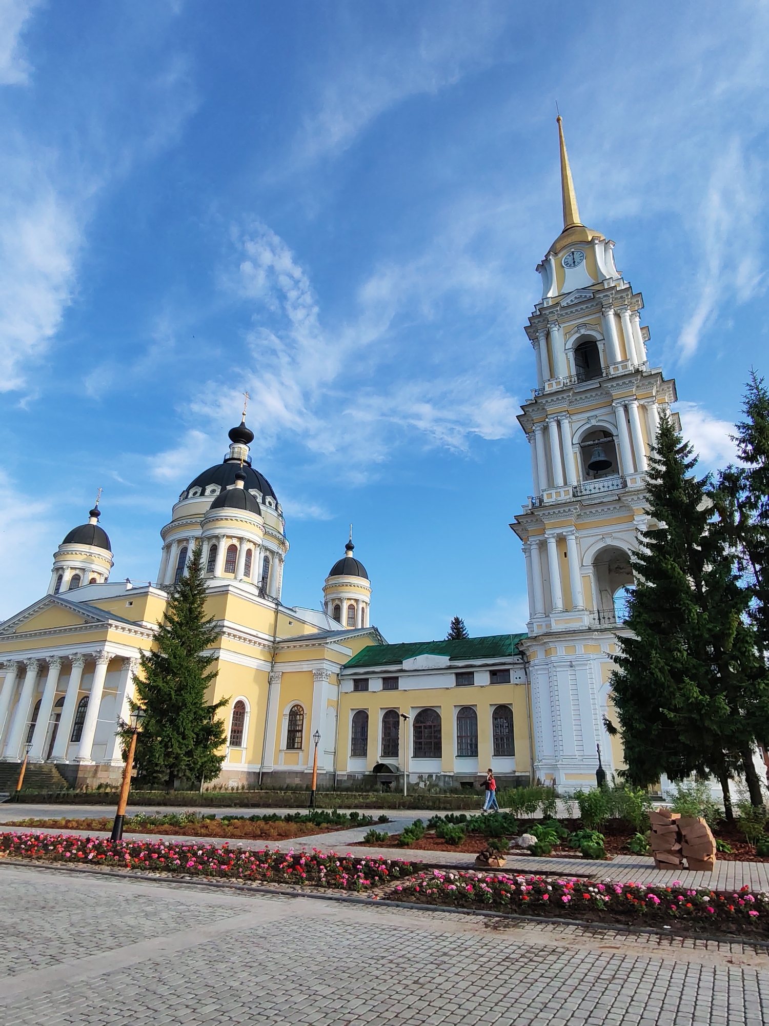 The image depicts the Church of the Transfiguration in Bol'shie Mkhi, Russia. The church features a prominent yellow facade with white architectural details, including columns and decorative elements. A tall bell tower with a golden spire stands to the right of the main church building, which has several black domes topped with crosses. The scene is set against a bright blue sky with scattered white clouds. In the foreground, a paved walkway leads to a garden area filled with blooming pink and red flowers. Several evergreen trees are planted around the church and in the garden. A single person, wearing a red jacket and blue pants, is walking on a pathway in front of the church.