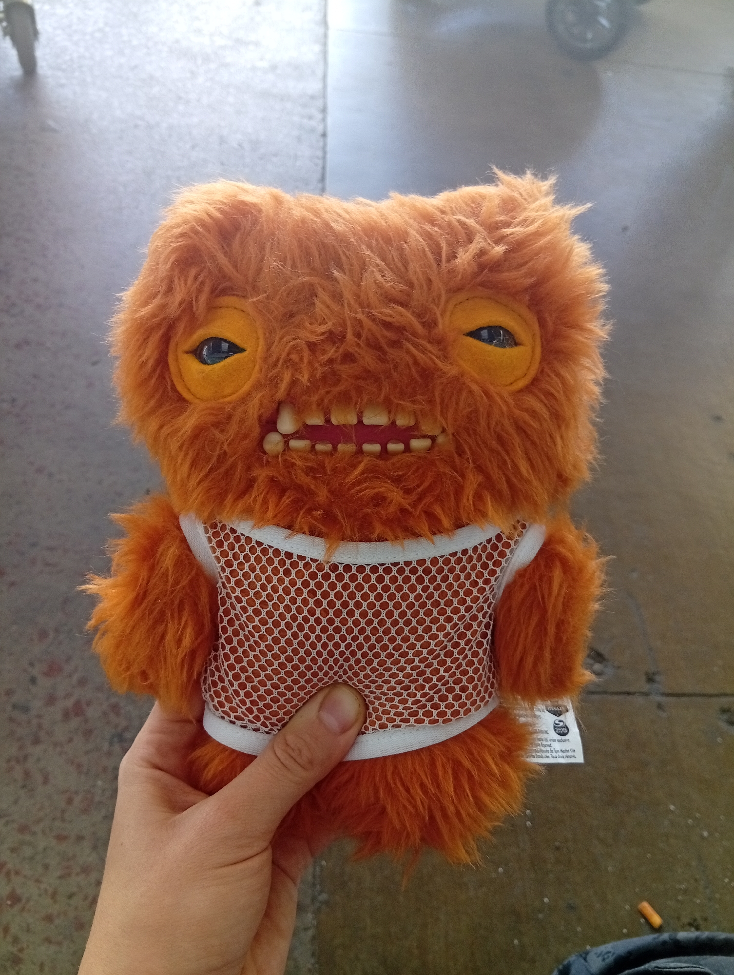 A person's hand holds an orange, furry plush toy with a grid-patterned vest against a blurred background. The toy has prominent teeth and yellow eyes with visible pupils. The background suggests an indoor setting, possibly a terminal, with floor tiles and vague shapes indicating distant objects. The location is Aeropuerto Internacional Ezeiza, Argentina.
