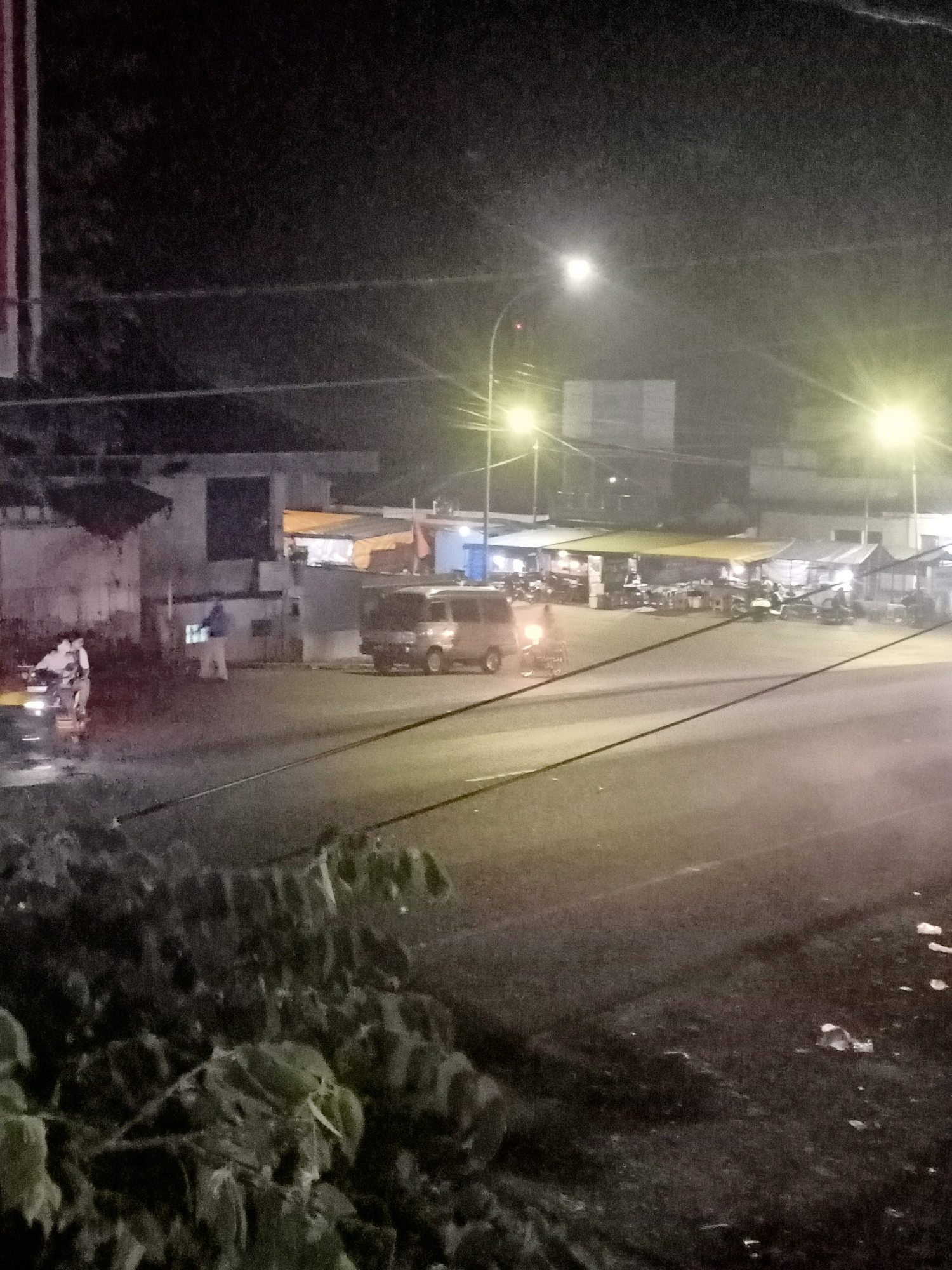 The image depicts a street scene at night in Jatisrono, Indonesia, characterized by artificial lighting from street lamps and illuminated stalls. On the left side of the road, two individuals are positioned on a motorcycle, appearing to be stationary. Nearby, a person in a light-colored shirt and pants stands beside a white object, possibly a cooler or a box. Further down the road, a light-colored van is parked. The background features several stalls or small shops with awnings, some of which are illuminated, suggesting commercial activity. The road surface is visible with clear lane markings, and the overall atmosphere is dim, with the lighting creating a hazy effect in some areas. Dense foliage is present in the foreground, obscuring part of the lower portion of the scene.