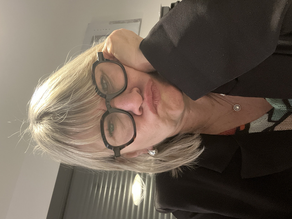 A blonde woman with glasses rests her head on her hand. She wears a black blazer over a patterned top and a delicate silver necklace. Her lips are pressed together in a neutral expression. The background includes a blurry light fixture and possibly a wall with vertical textures.